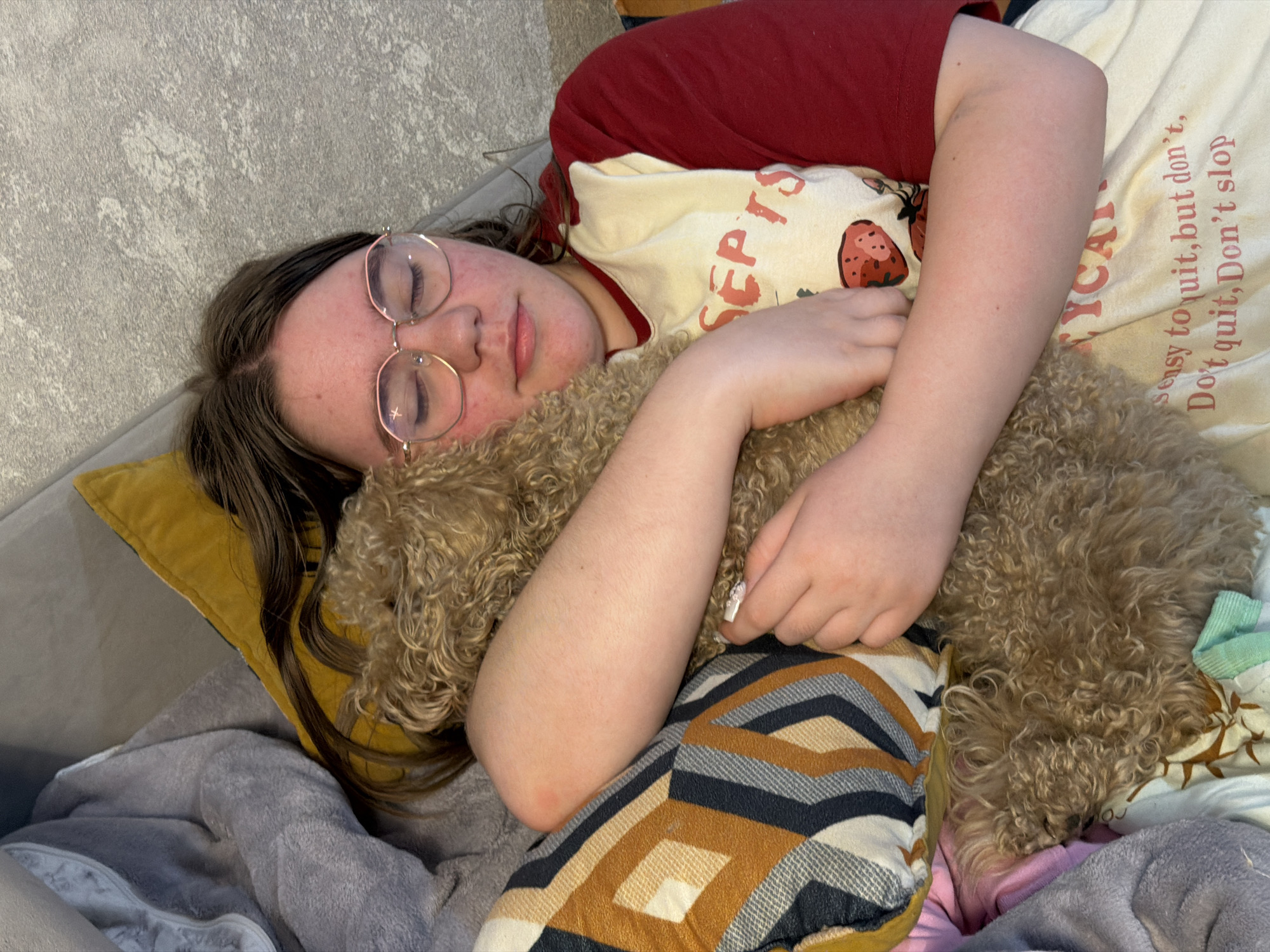 In the city of Perm, Russia, a person is sleeping on a couch, embracing a small, light brown poodle. The person, wearing glasses and a t-shirt with a strawberry print and text, is lying on their side, with their head resting on a yellow pillow and a gray blanket. The poodle is nestled close to the person, partially obscured by their arm. The background shows a textured wall and other pillows and blankets. The lighting is soft, suggesting an indoor setting.