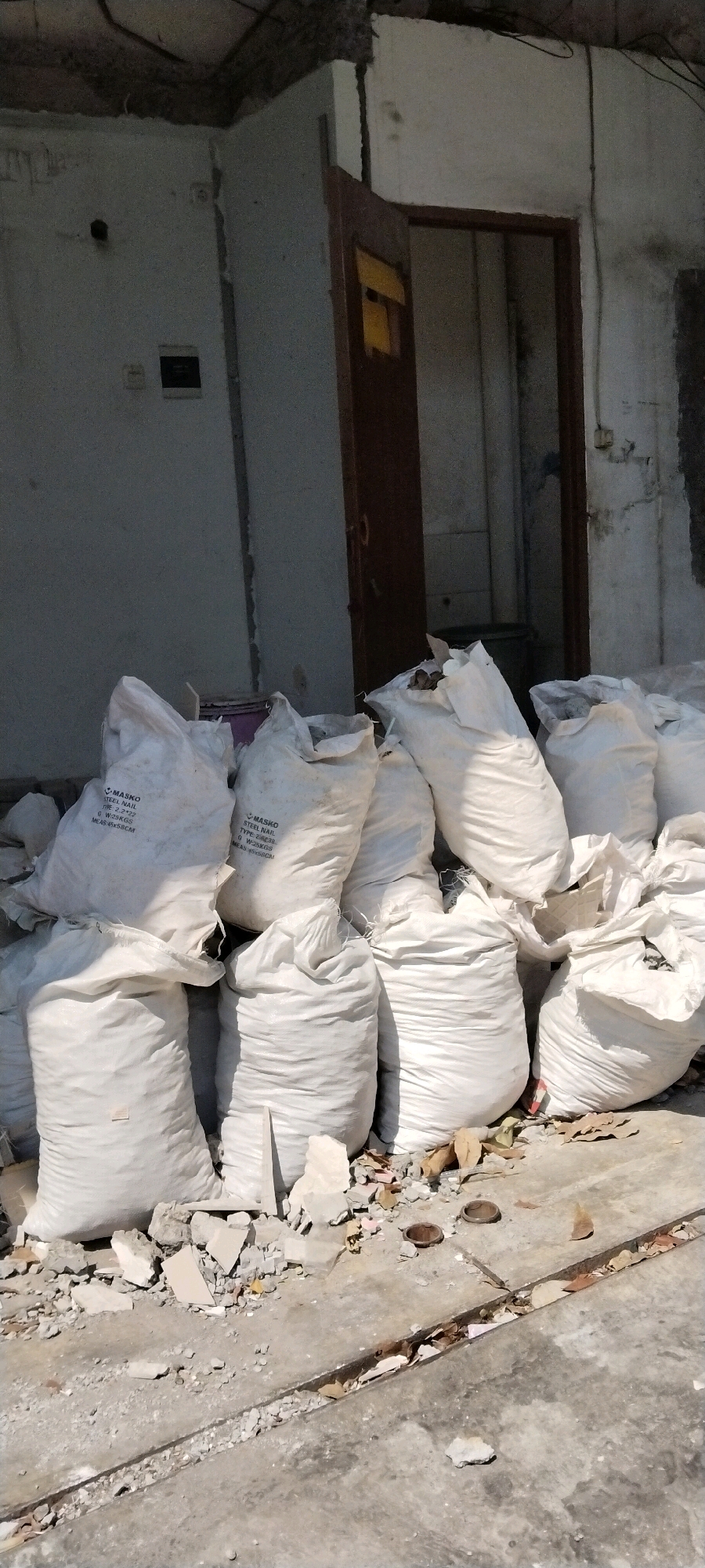 A pile of large, white, burlap sacks, likely filled with construction debris, is staged on a concrete surface. The sacks are of varying sizes and are haphazardly stacked. Some of the sacks have text printed on them, indicating they contain "MASKO STEEL NAIL." The ground around the sacks is littered with broken concrete, debris, and dry leaves. In the background, a doorway with a partially open wooden door leads into a dark interior. The wall behind the sacks is weathered and appears to be part of an unfinished or damaged building. The overall scene suggests an outdoor construction or demolition site in an urban environment.