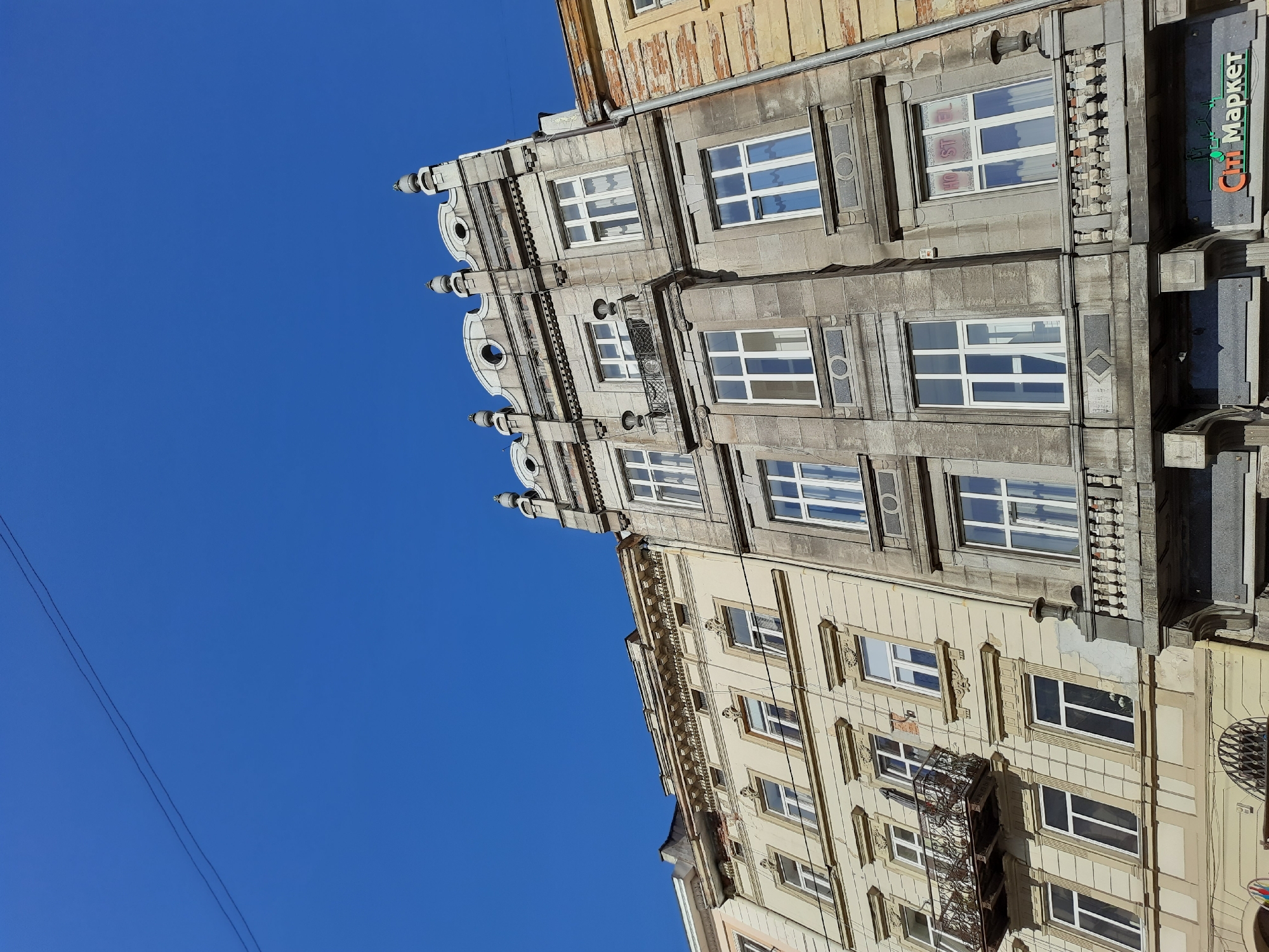 This is a low-angle, upward shot of several old buildings in the city of Lviv, Ukraine, under a clear blue sky. The buildings are constructed in a European architectural style, with multiple stories, ornate facades, and numerous windows. The building on the right is made of stone and features decorative elements like balustrades and sculpted pinnacles. The building to its left is lighter in color, possibly stucco, and also displays decorative window frames and balconies. A black wire runs diagonally across the upper part of the image, with another thinner wire running horizontally across the facades of the buildings. Some of the windows have white curtains, while others show reflections. On the ground level of the stone building, a sign with green lettering reads "Ситі Маркет," indicating a market or store. The overall impression is one of historic urban architecture bathed in sunlight.