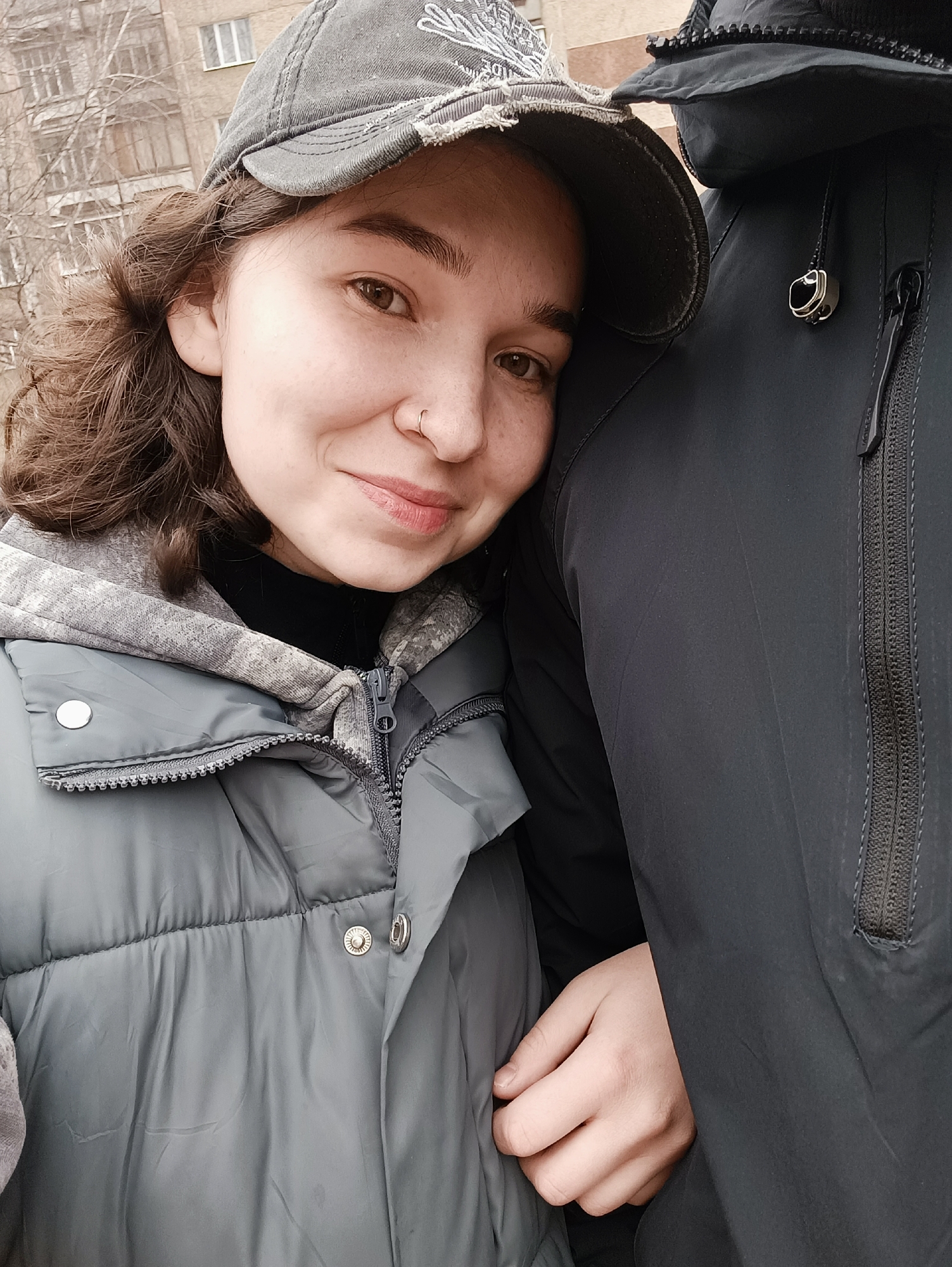 A close-up shot captures a woman with fair skin, brown curly hair, and a nose ring. She wears a gray cap and a gray hooded jacket. The jacket is partially zipped and appears to be a quilted, padded style.
The woman is leaning against a person wearing a dark jacket, their arm around her. Only the person's torso, shoulder, and part of their hand are visible.
In the background, a multi-story building can be seen. Bare tree branches are visible in front of the building.
Given the information available, the exact location in Izhevsk, Russia, cannot be determined with certainty. However, the presence of bare trees and overcast lighting suggests an urban environment and possibly a colder season.