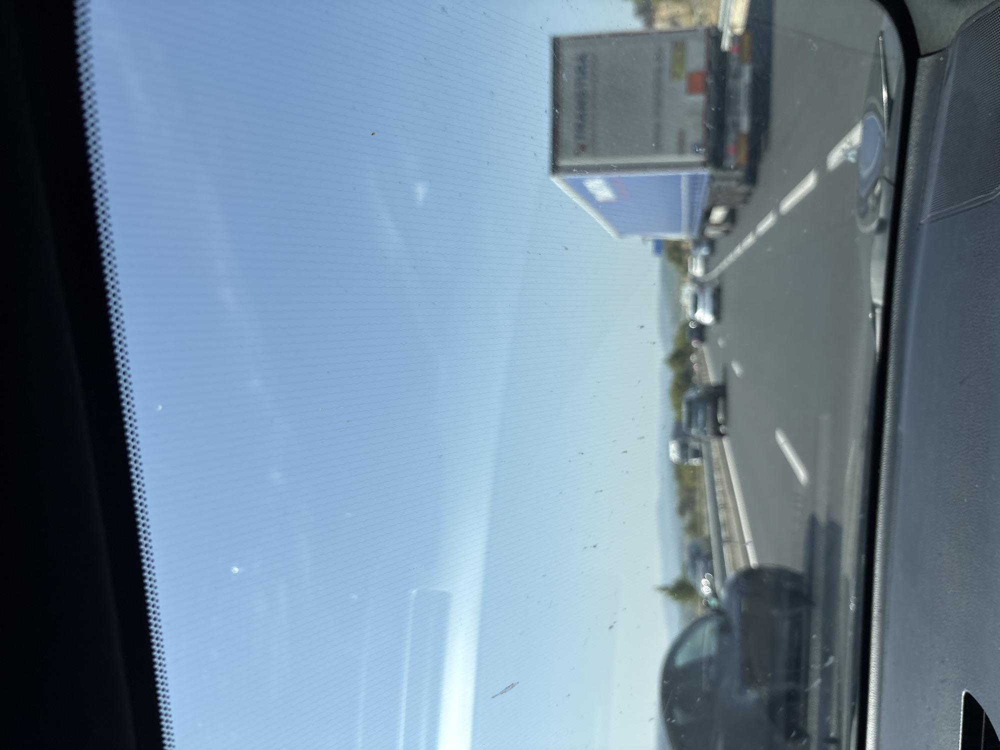 The image shows a view from the windshield of a car driving on a highway. The sky is blue with some wispy clouds. There are several cars and a large blue and white truck on the road. The highway appears to be in a rural or semi-rural area, with hills visible in the distance. The car is in motion, as indicated by the blurred foreground and the perspective of the road. There are some smudges and dust on the windshield. There is no indication of the location being specifically within the city of Bages, France.