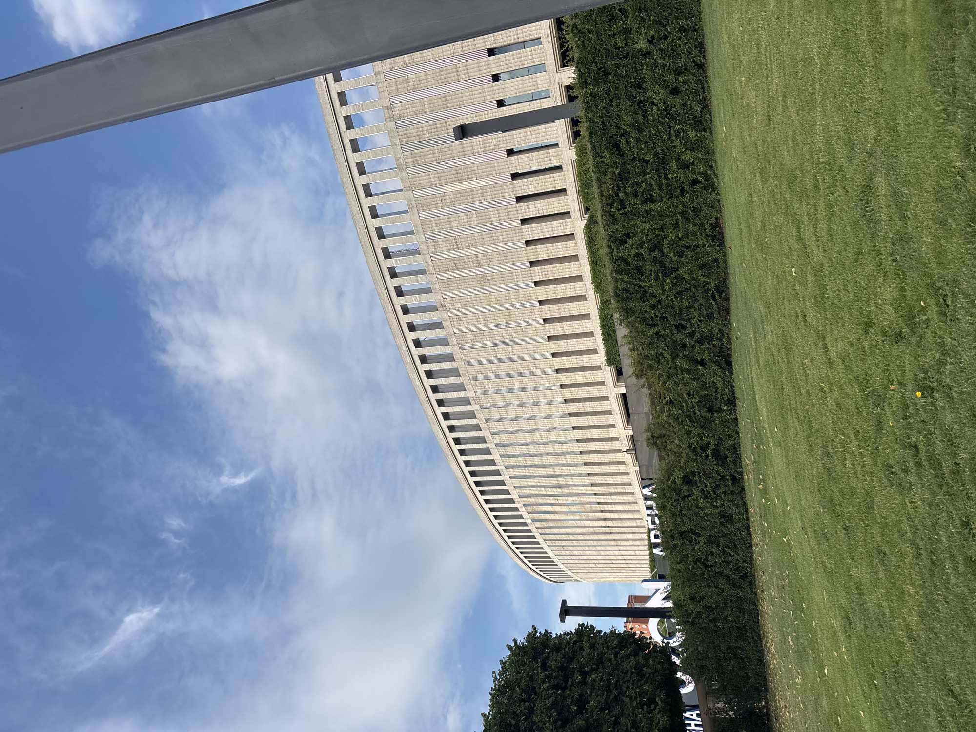 This media file depicts an outdoor scene with a large, modern building and a grassy area. The building, which appears to be a stadium or arena due to the curved architecture and large windows, is a prominent subject. It is constructed with light-colored panels and features numerous rectangular openings. Adjacent to the building is a dense, green hedge that forms a boundary. Beyond the hedge, a well-maintained lawn stretches out, with some scattered yellow leaves. The sky above is blue with scattered white clouds, suggesting a pleasant day.
The image's orientation is rotated, with the top of the frame appearing on the left. A large, grey metal beam runs diagonally across the upper left portion of the image, possibly part of an overhead walkway or structure. On the building's facade, a white sign with dark lettering is partially visible, reading "ARENA" and "CHA" with the letters "N" and "O" also discernible, hinting at a location name. A dark, slender pillar stands near the building, casting a shadow. Below this, the top of a dark green tree is visible.
The time of day appears to be daytime, likely late morning or early afternoon, given the bright lighting and clear sky. The weather seems to be mild and sunny. The scene is set in an urban or semi-urban environment, possibly a sports complex or public venue, given the "ARENA" signage. There are no people visible in the image, and no specific activity or event is taking place; it is a static depiction of the location. The overall mood conveyed is one of modern architecture and well-kept grounds.