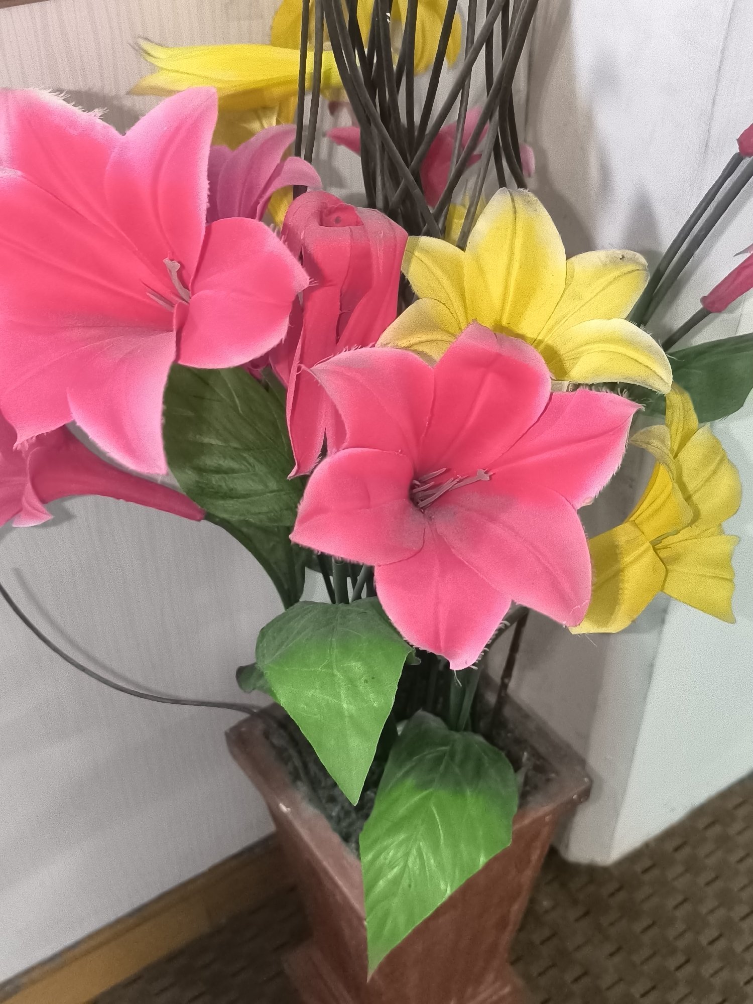 This is a close-up, eye-level, indoor photograph of a bouquet of artificial flowers in a planter. The bouquet features several large, vibrant pink lily-like flowers and some yellow lily-like flowers, along with green artificial leaves. The flowers appear to be made of fabric and have a slightly fuzzy texture, with some dust visible on the petals. Dark, thin stems rise from the arrangement, some topped with what appear to be additional pink flower buds. The planter itself is a reddish-brown, rectangular pot with a textured surface. In the background, a lightly textured, pale wall is visible, and a wooden surface with a woven pattern runs along the bottom of the frame. The lighting is soft, suggesting an indoor setting. There are no people or animals visible in the image. This scene is likely found within a home or a commercial establishment in Kalabbirang, Indonesia, given the provided context.