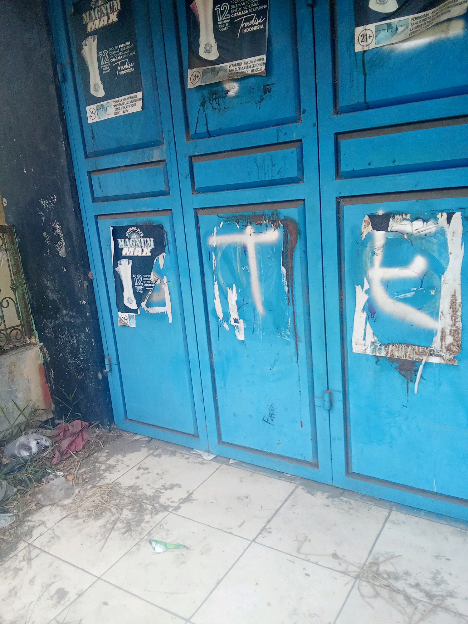 The image depicts a weathered, blue metal door in Warunggunung, Indonesia, likely a shop front. The door is divided into two main panels, with a vertical seam running down the center. Both panels are adorned with various posters and graffiti.
On the left panel, a torn poster for "MAGNUM MAX" cigarettes is visible, featuring an image of a cigarette pack and warning labels. Below it, a smaller circular sticker with "(21+)" is affixed. The door itself shows signs of wear and tear, with peeling paint and rust streaks.
The right panel of the door is covered in large, white graffiti that resembles stylized letters or abstract shapes. Remnants of torn posters are also visible underneath the graffiti. A "(21+)" sticker is present on this panel as well.
The ground in front of the door is paved with light-colored tiles. Debris, including dried leaves, grass, and what appears to be discarded plastic packaging, is scattered across the ground and near the base of the door. Some of this debris is also visible in the lower left corner of the image, including a crumpled plastic bag and a piece of red fabric. The overall scene suggests an urban environment that is somewhat neglected.