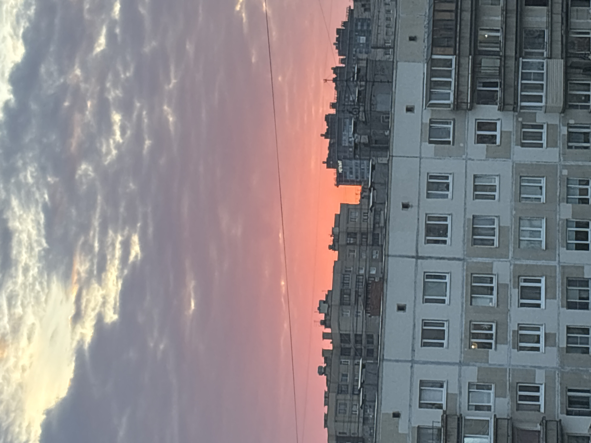 The image, oriented vertically, captures an urban landscape at either sunrise or sunset. The sky, occupying the upper left portion of the frame, displays a striking gradient of colors. On the far left, light grey and white clouds are prominent, reflecting a bright, possibly early morning or late evening sun. These clouds transition into a soft purple-pink hue as they stretch towards the horizon, eventually meeting a vibrant, clear expanse of pink and orange sky directly above the city skyline.
A large, multi-story apartment building dominates the right side of the frame. Its facade is composed of light grey or beige panels, interspersed with darker grey or patched sections, indicating some wear or renovation. Numerous square windows, many with visible frames and some with reflections, are systematically arranged across its surface.
Below the colorful horizon, a dense silhouette of a city skyline is visible, consisting of various buildings of different heights and architectural styles, suggesting a bustling urban environment. A thin, dark power line or cable stretches horizontally across the middle of the image, cutting through the gradient of the sky, connecting unseen points.
No people or discernible activities are visible, emphasizing the static nature of the urban vista. The overall mood conveyed is tranquil and contemplative, marked by the dramatic natural light interacting with the structured geometry of the city. The architectural style of the buildings suggests a potential Eastern European or post-Soviet setting. There is no clearly legible text visible in the image.
