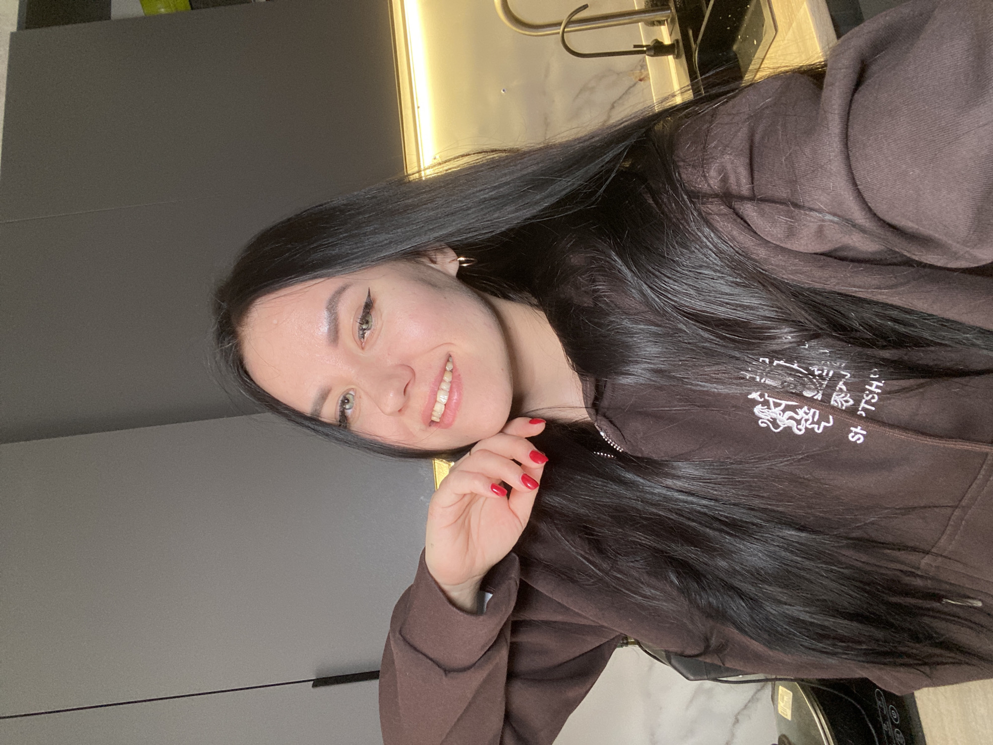 A young woman is captured in a diagonal selfie, smiling broadly and looking directly at the camera. She has long, dark, straight hair that frames her face and extends over her shoulders and chest. Her eyes are light-colored, and she wears noticeable black winged eyeliner. Her smile reveals her teeth, conveying a cheerful and happy demeanor. Her right hand, with bright red nail polish on her fingernails, is held gently near her face.
She is wearing a brown zippered hoodie. White text and graphics are partially visible on the hoodie, with discernible letters forming "SPORTSH." and an intricate, stylized design, possibly a crest or an animal motif.
The background suggests an indoor setting, likely a modern kitchen or apartment in Chaikovsky, Russia. To the left, sleek, dark gray cabinets are visible. In the upper right corner, a kitchen faucet with a metallic finish and a light-colored, possibly marble-patterned countertop can be seen. A warm, artificial strip light illuminates the area under an overhead cabinet, casting a glow on the background elements. The overall impression is one of a casual, engaging portrait within a contemporary home environment.