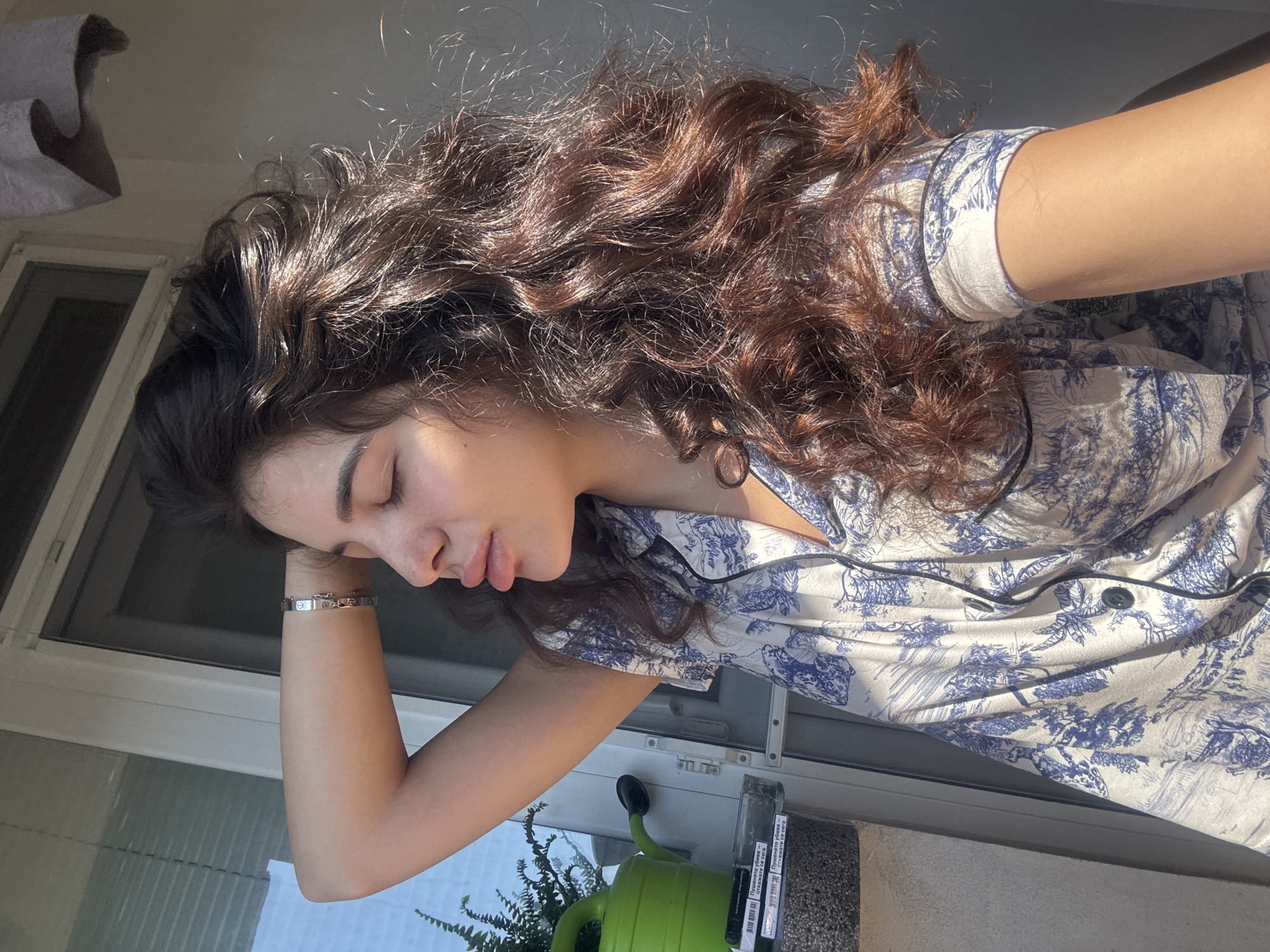 The image shows a young woman with long, wavy, dark brown hair lying down with her eyes closed. She is wearing a light-colored pajama top with a blue toile pattern. Her right arm is bent, and she is resting her head on her hand, wearing a silver bracelet on her wrist. The lighting suggests it is daytime, possibly late afternoon, with sunlight illuminating her hair and skin. The background shows a window with blinds and a view of what appears to be an outdoor balcony or ledge. A green watering can and a plant are visible in the lower right corner. Two labels with Cyrillic text are attached to the watering can. The overall mood is peaceful and relaxed. The location context is Plovdiv, Bulgaria.