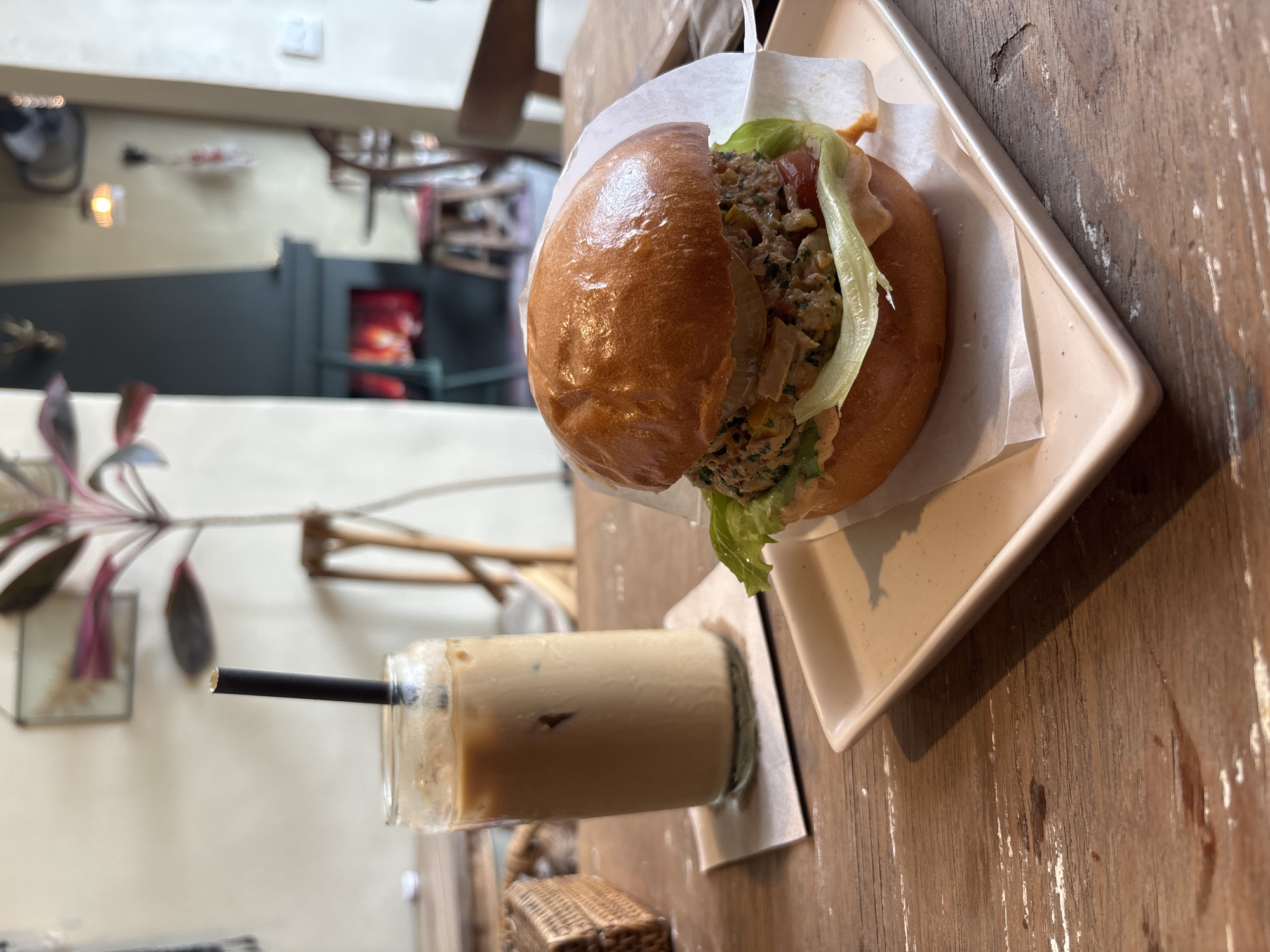 The image depicts a meal on a wooden table. A burger on a square plate, lined with white paper, takes center stage. The burger appears to contain lettuce and other fillings. Next to the burger, is a glass of iced coffee with a black straw. The background shows a glimpse of an interior space, with a plant, wooden furniture, and a partially visible room with a light fixture. Given the prompt's request to specify the location in Kuta Selatan, Indonesia, it can be deduced that the photograph was taken in a restaurant in Kuta Selatan. The scene seems to be a casual dining setting, possibly during lunch or brunch.
