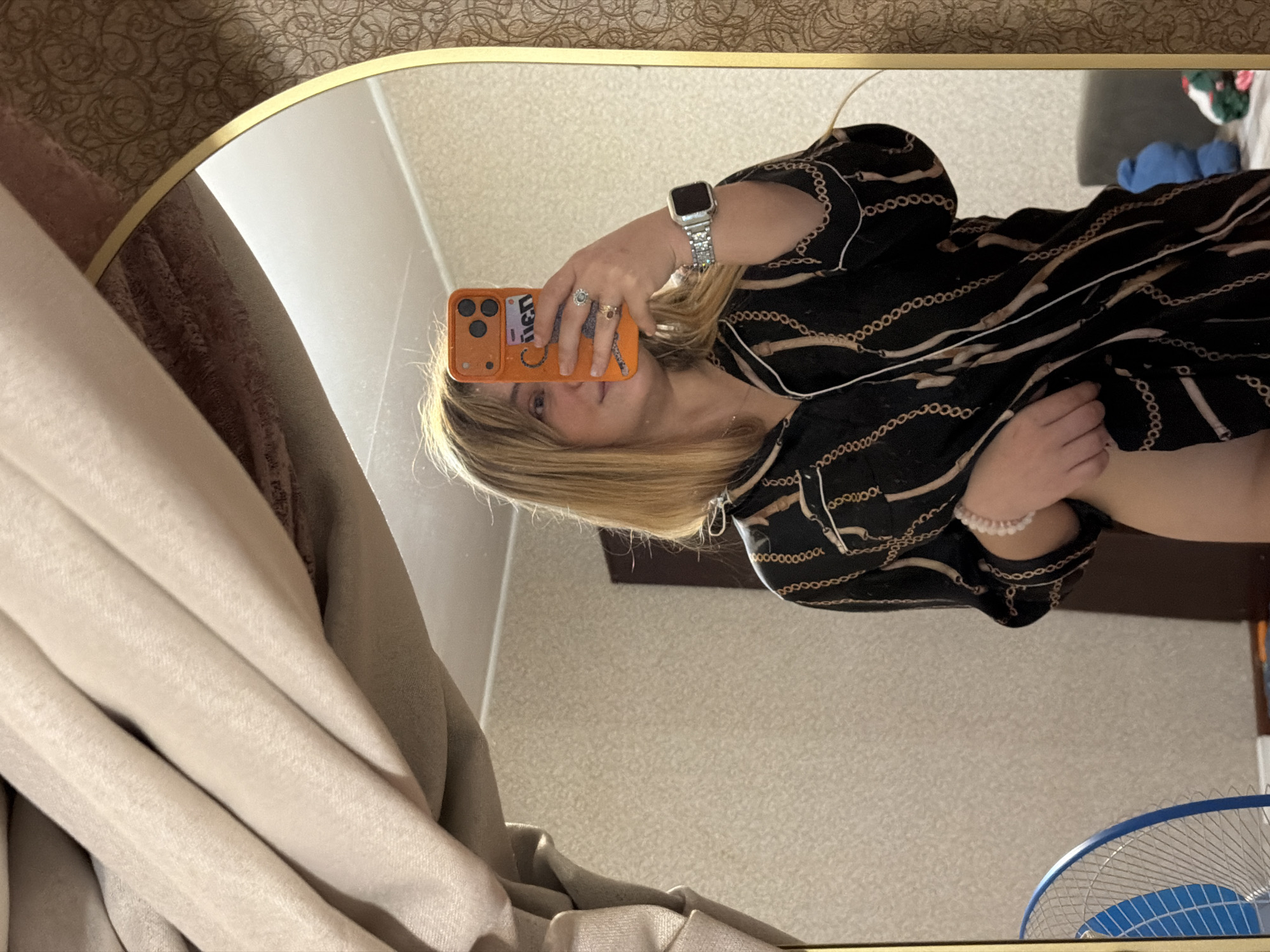 The image is a selfie taken in a mirror. The reflection shows a woman with blond hair holding an orange iPhone. She is wearing a black and tan patterned blouse. On her left wrist, she has a silver-toned smartwatch and a pink beaded bracelet. Multiple rings adorn her fingers. The background reveals a beige wall, a curtain on the left, and what appears to be a blue-and-white fan in the lower right corner. The location is identified as Naberezhnye Chelny, Russia.