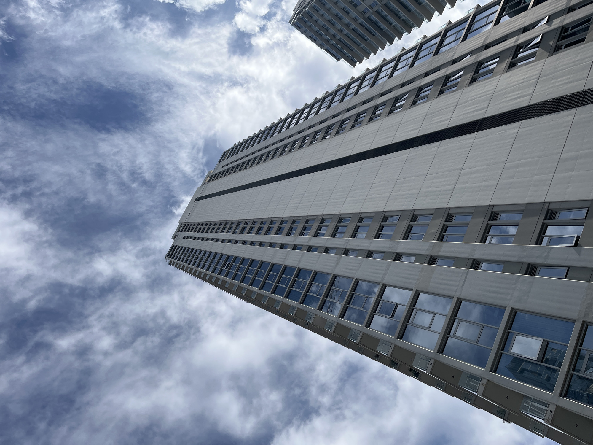 The image shows a worm's-eye view of a tall, modern building with a gray facade and numerous windows that reflect the cloudy sky. The building has a simple rectangular shape with a dark horizontal line running across its middle section. A portion of another similar building is visible in the upper part of the frame. The sky is filled with a mix of white and gray clouds. The exact location cannot be determined from the image. The city of Buenos Aires, Argentina cannot be verified from this image.