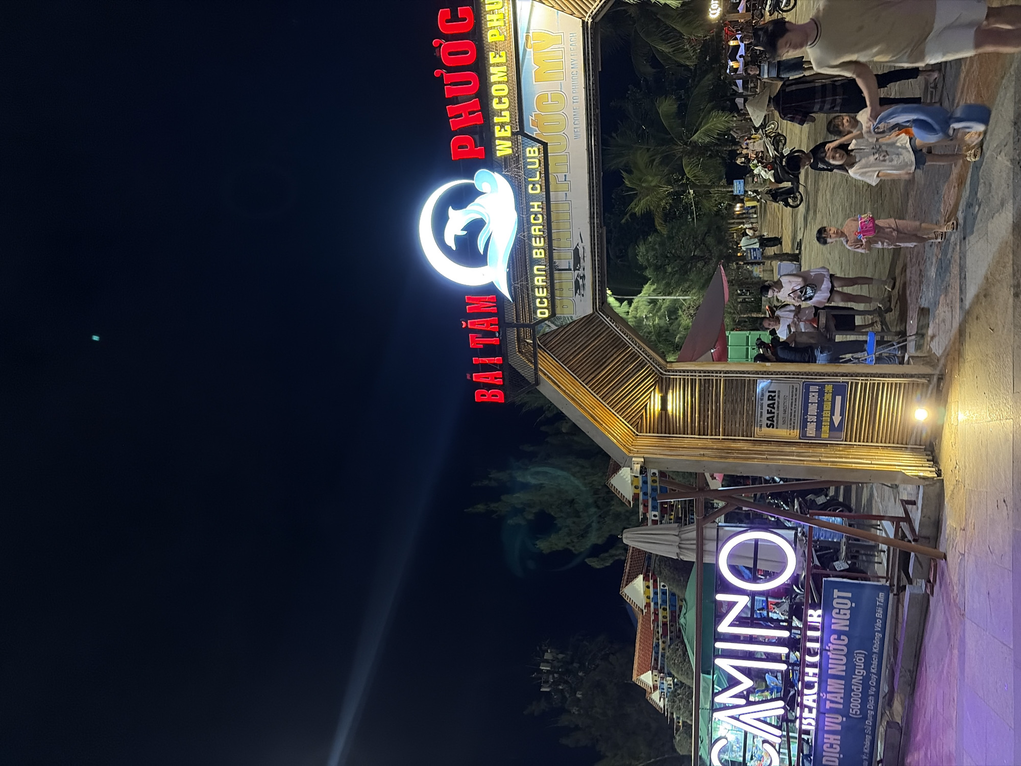 The image shows a neon-lit entrance to a beach club at night. The main sign reads "BÃI TẮM PHƯỚC MỴ OCEAN BEACH CLUB" with a dolphin logo. To the left, another sign displays "CAMINO BEACH CLUB" in neon. A blue banner underneath that reads "DỊCH VỤ TẮM NƯỚC NGỌT (5000₫/Người)" which translates to "FRESHWATER BATHING SERVICE (5000₫/Person)". The entrance is framed by a bamboo structure. In the background, palm trees and other buildings are visible, along with several people walking and standing around. Motorcycles are parked near the entrance. The overall scene suggests a lively tourist area.