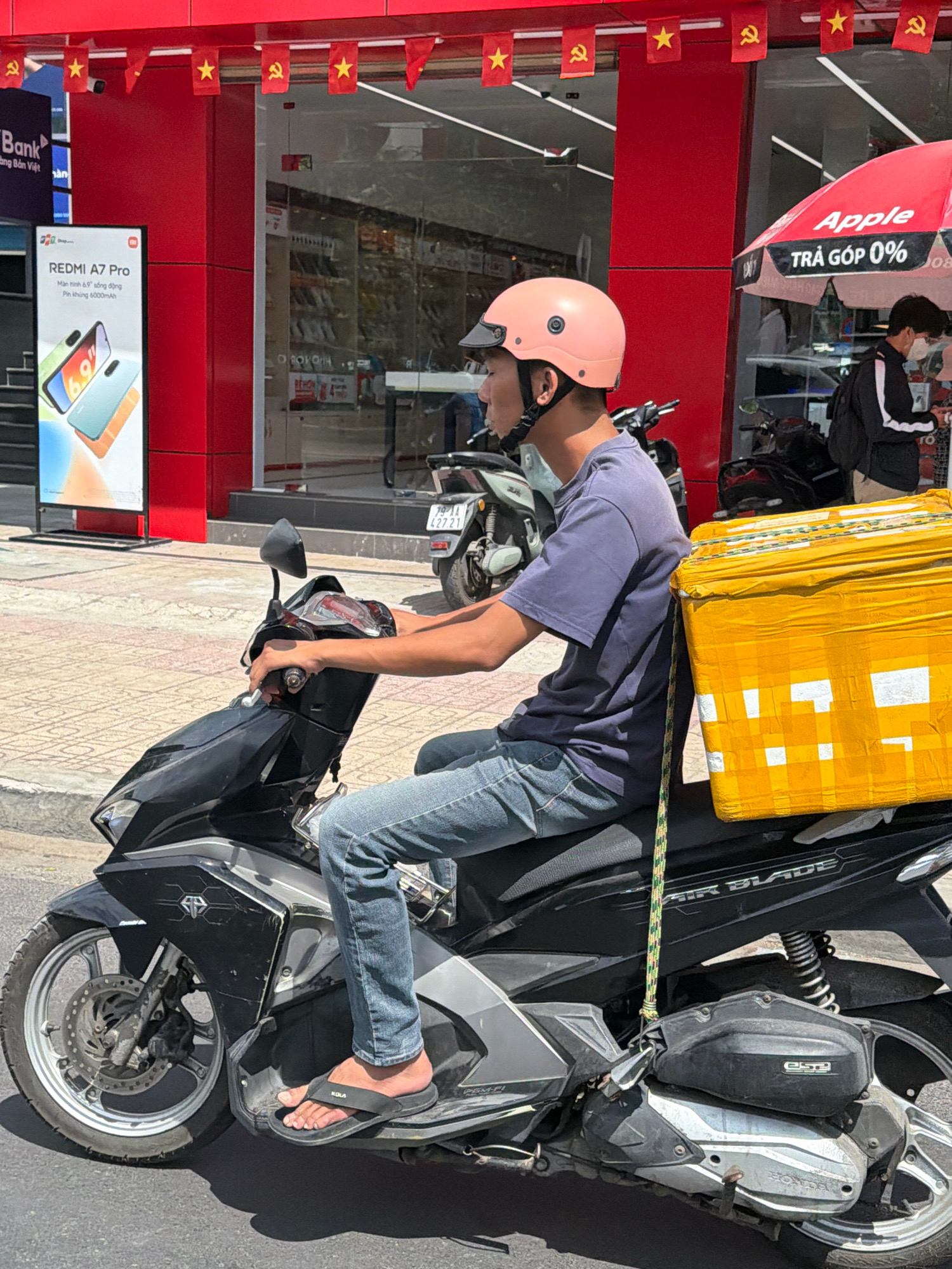 A young man wearing a pink helmet and blue jeans rides a black Honda Air Blade scooter. He is carrying a large yellow box secured to the back of the scooter with a rope. The scooter is parked on a paved street in front of a red building with red banners hanging overhead. The building appears to be a store, with a large window displaying merchandise. There is an open red umbrella with "Apple" and "TRẢ GÓP 0%" written on it in white letters to the right of the scooter. The scene is set on a sunny day. The image appears to have been taken in Nha Trang, Vietnam, given the presence of Vietnamese text on the umbrella and the general appearance of the street and buildings.