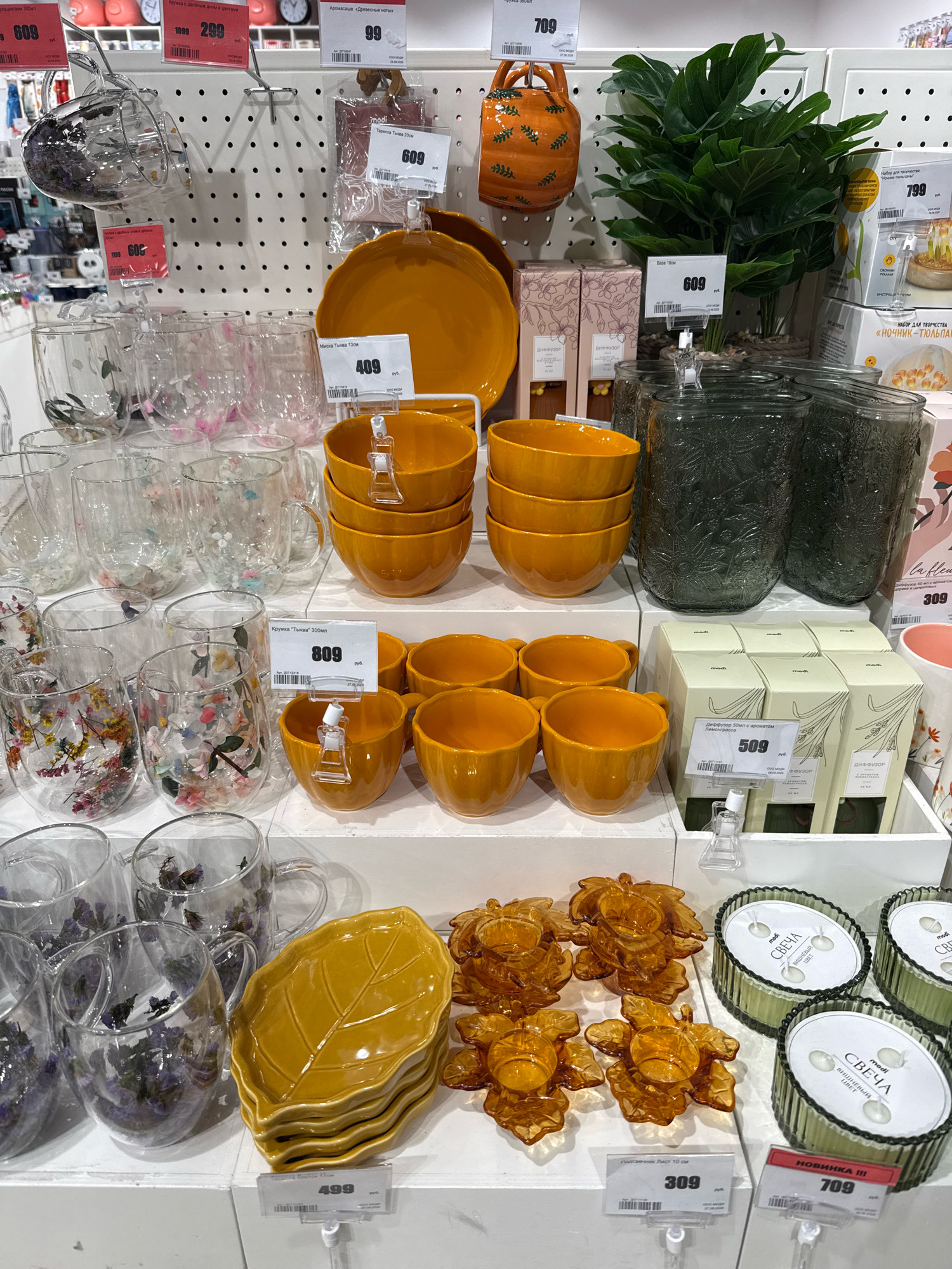 The image displays a retail shelf in a store in Bor, Russia, featuring a variety of home goods. The products visible include a collection of clear glass mugs and cups, some decorated with floral or botanical patterns, and others containing dried flowers or herbs. There are also stacks of yellow, leaf-shaped ceramic plates and several amber-colored glass candle holders shaped like leaves. Additionally, large dark green glass jars with embossed patterns, and boxes of diffusers are present. Price tags are attached to many of the items, indicating prices in rubles. The lighting suggests an indoor environment typical of a retail store. No people are visible in the image.