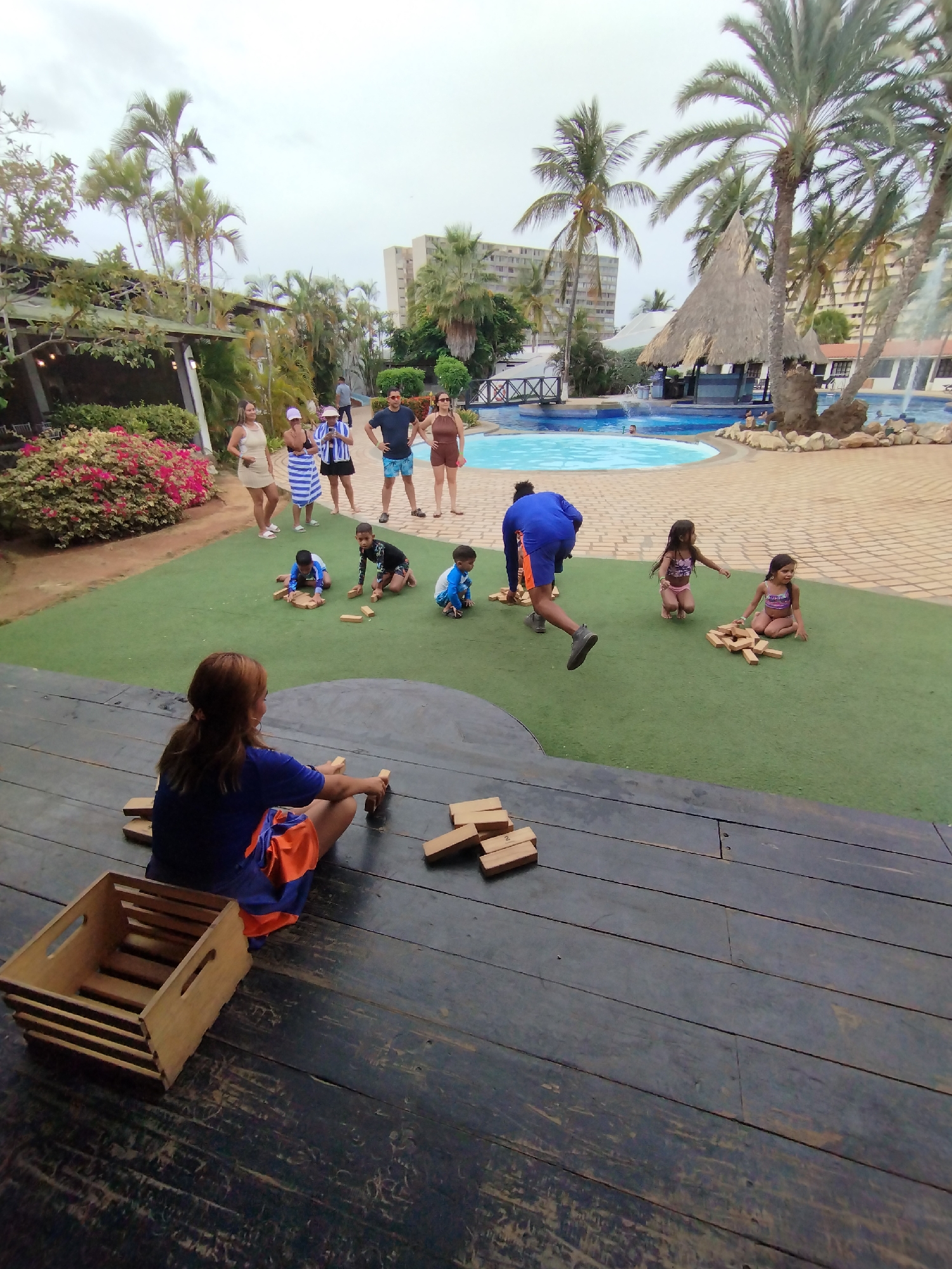 The image captures an outdoor leisure scene, seemingly at a resort or hotel in Porlamar, Venezuela, given the presence of palm trees, a swimming pool, and buildings in the background. In the foreground, a woman is seated on a weathered, dark wooden platform, arranging wooden blocks, possibly for a game of Jenga, with an empty wooden crate next to her. On a patch of artificial turf above the platform, several children are engaged in playing with similar wooden blocks, while a man in a blue outfit appears to be in motion, stepping away from the children. A group of adults stands further back near the pool area, observing the children. The sky is overcast, suggesting a cloudy day. The scene suggests a family-friendly environment with recreational activities.
