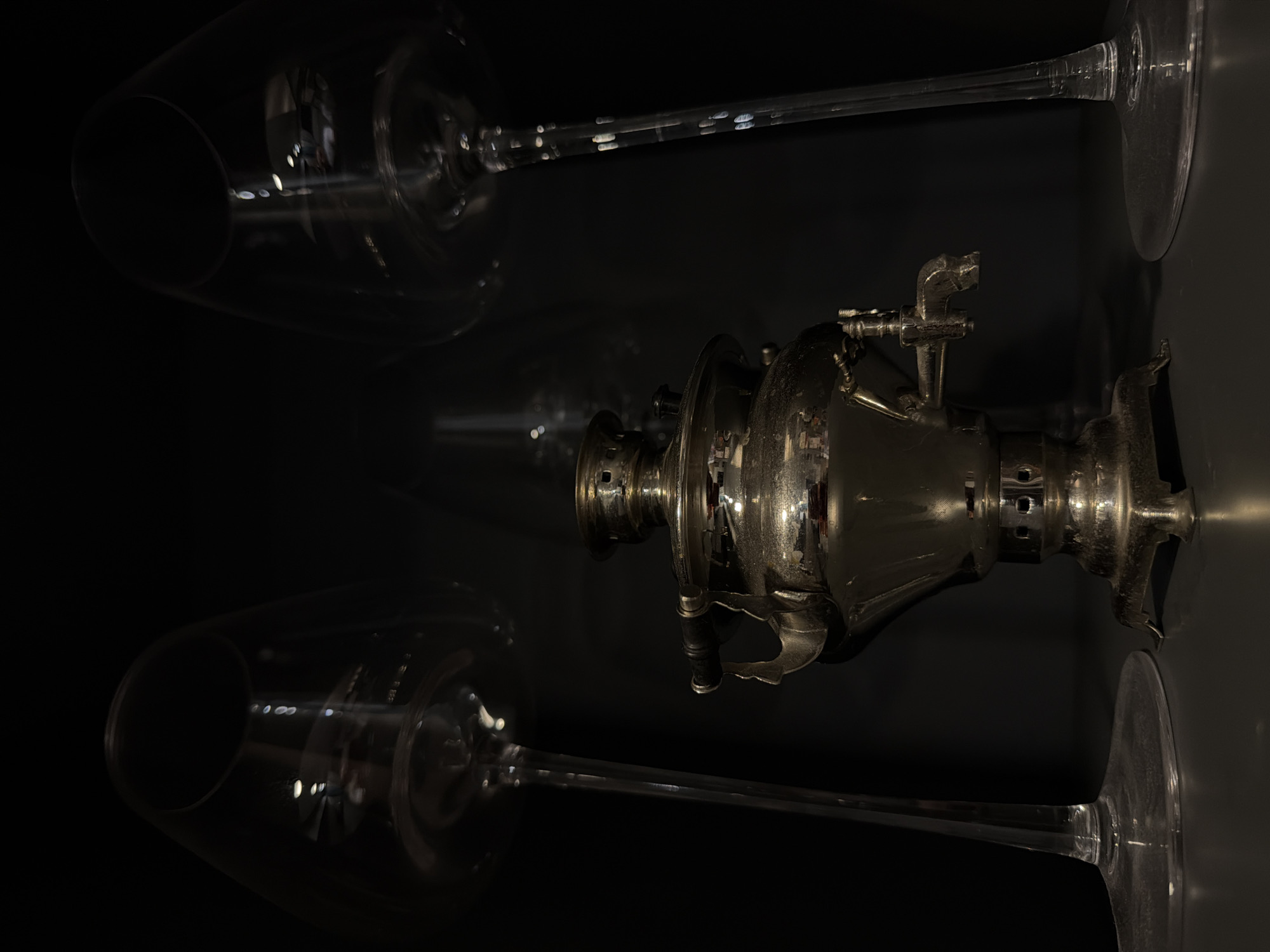 The image presents a still-life composition in a dark environment. The primary object is a metallic samovar, positioned centrally in the frame. Two wine glasses flank the samovar, one above and one below, appearing to be in a state of being laid down. The glasses are transparent, and their stems are visible. The scene exhibits significant contrast, with the metallic objects reflecting light in an otherwise dark setting. There is no information that this image was taken in Birobidzhan, Russia.