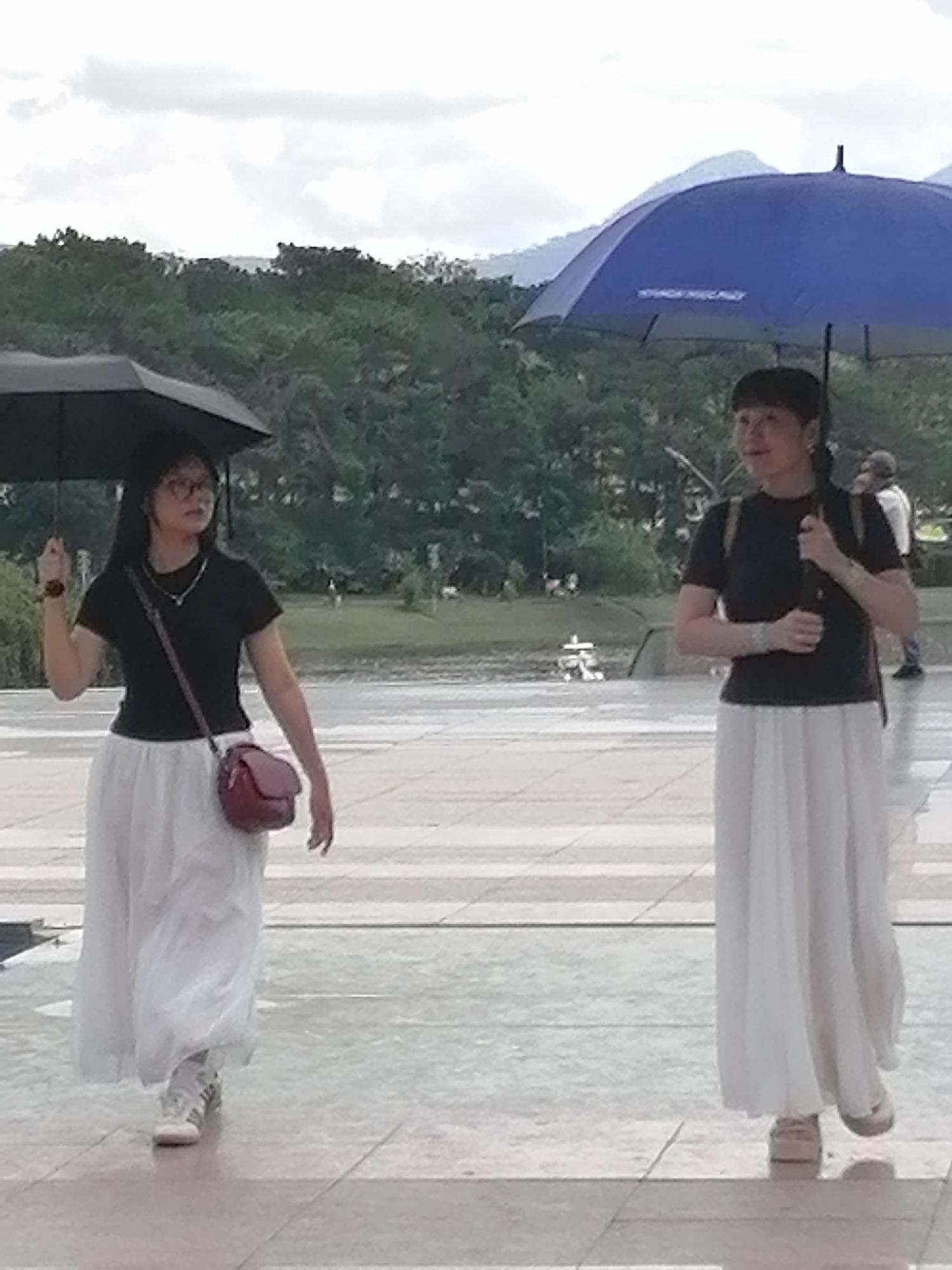 Two individuals walk outdoors, each holding an umbrella. The person on the left, a female, is wearing a black t-shirt, a white flowing skirt, glasses, and carries a maroon crossbody bag. She is holding a black umbrella. The person on the right, also female, is wearing a black t-shirt and a white flowing skirt. She is holding a blue umbrella. They are walking on a paved walkway. In the background, there is a body of water, trees, and hills. The overall setting appears to be a park or scenic area, potentially in Da Lat, Vietnam, given the lush greenery and hilly landscape often found in the region. The weather seems overcast.