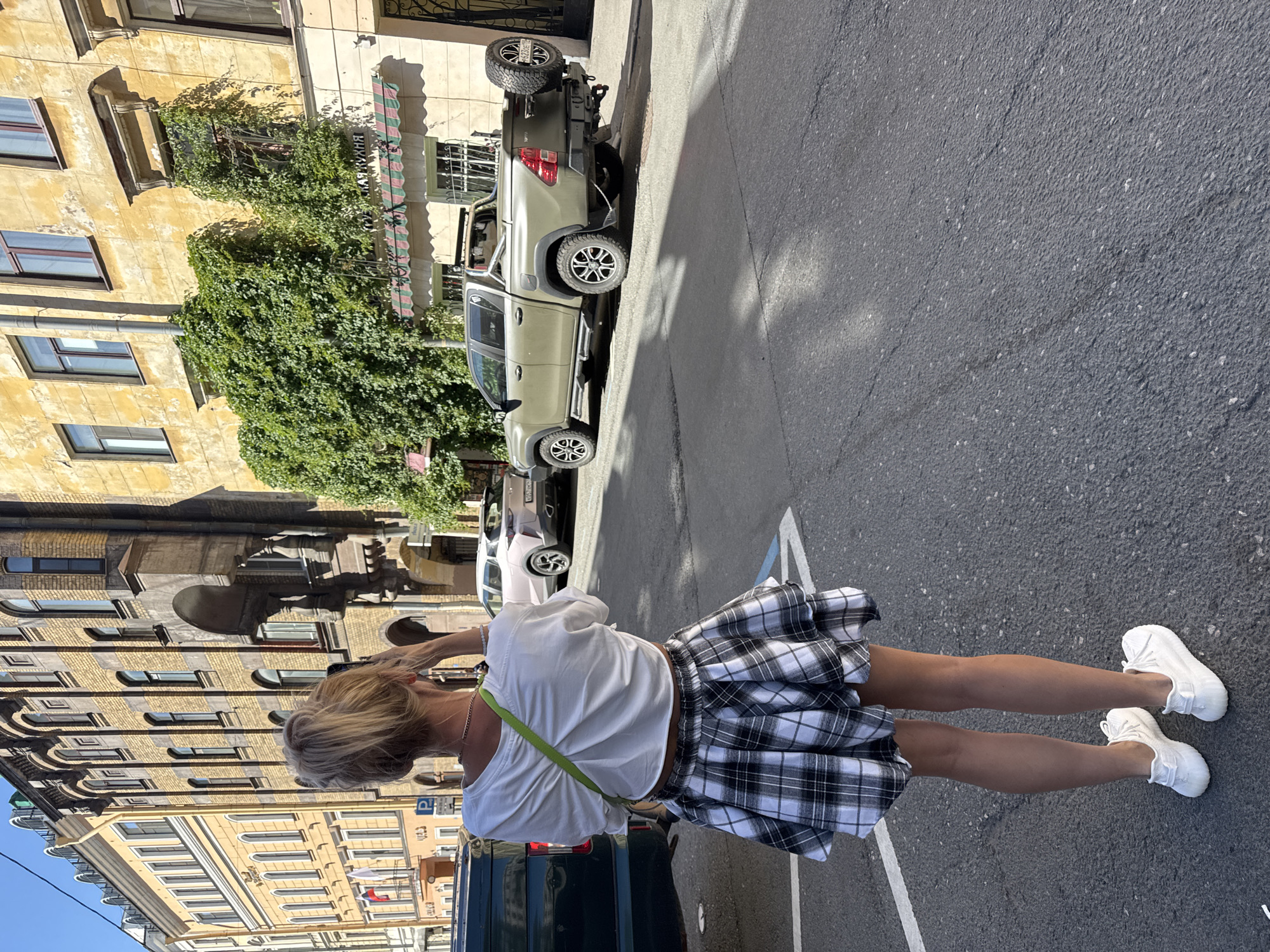 In Saint Petersburg, Russia, a woman stands on a paved city street, facing away from the camera. She is dressed in a white t-shirt, a black and white plaid skirt, and white sneakers. She holds a mobile phone up to her face, appearing to take a photograph. Her blonde hair is pulled back in a bun, and she wears a green strap across her chest, likely from a bag. The street is bordered by old European-style buildings with yellow facades. A dark green van is parked on the left side of the frame, and a khaki pickup truck with a spare tire mounted on the back is parked on the right. The sky is clear and blue.