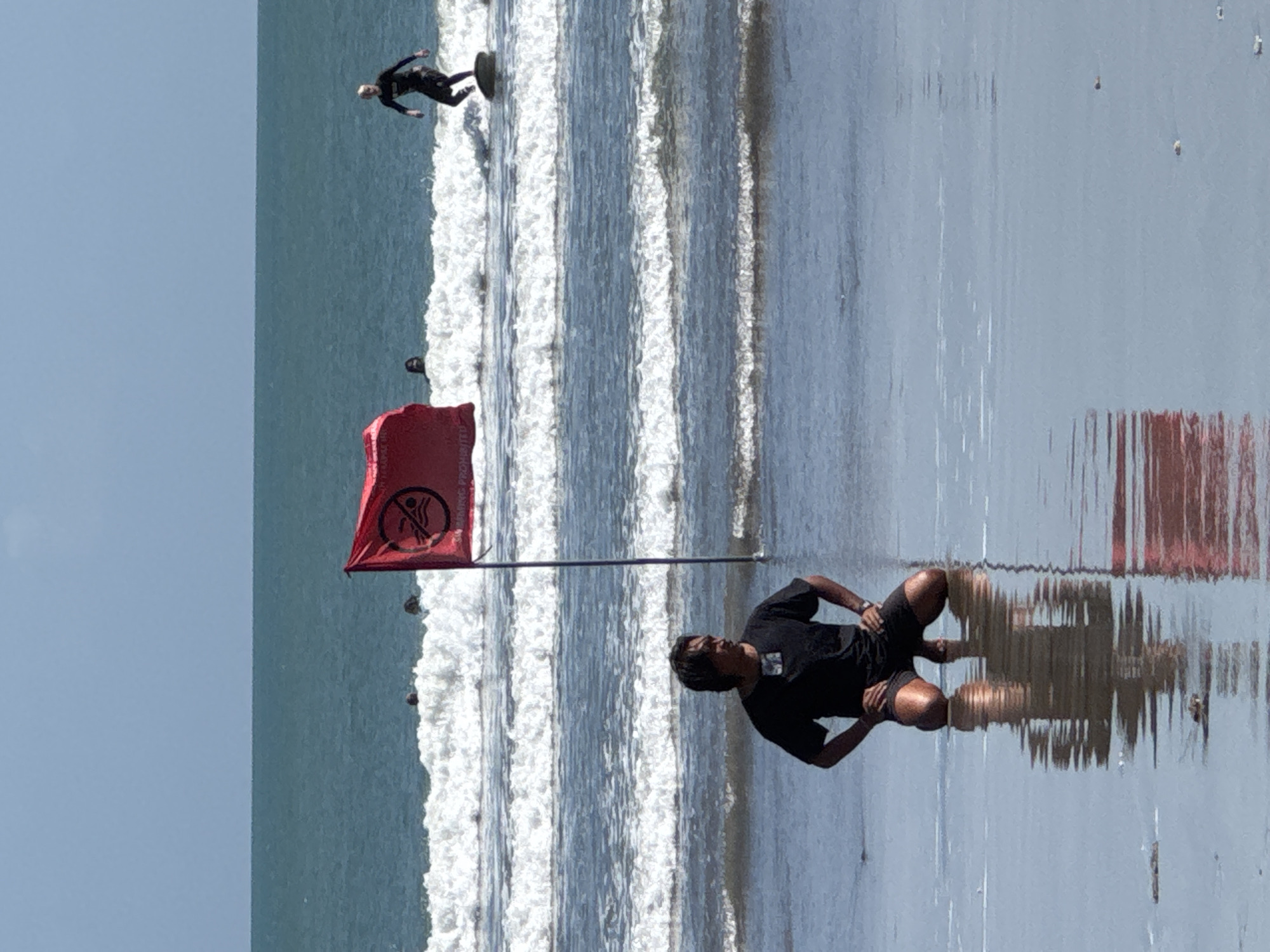 At Kuta Beach, Bali, Indonesia, a man in black shorts and a black t-shirt kneels on the wet sand. His reflection is visible in the shallow water. A red flag with a "no swimming" symbol is planted in the sand behind him. In the ocean, waves break, and a surfer in a black wetsuit rides a board to the right. Several other figures are visible in the water. The sky is clear and blue, and the sea is a deep blue-green.