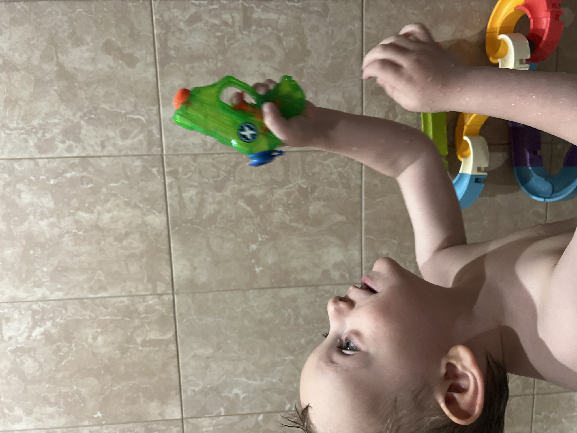 A young child is in a bathroom with tiled walls. They are holding a green and blue water gun in their right hand, pointing it upwards. A colorful toy consisting of interconnected circular and curved pieces is affixed to the wall behind the child's arm. The child is looking upwards, and their skin appears wet. Location: Unascertainable, though Mytishchi, Russia was specified.