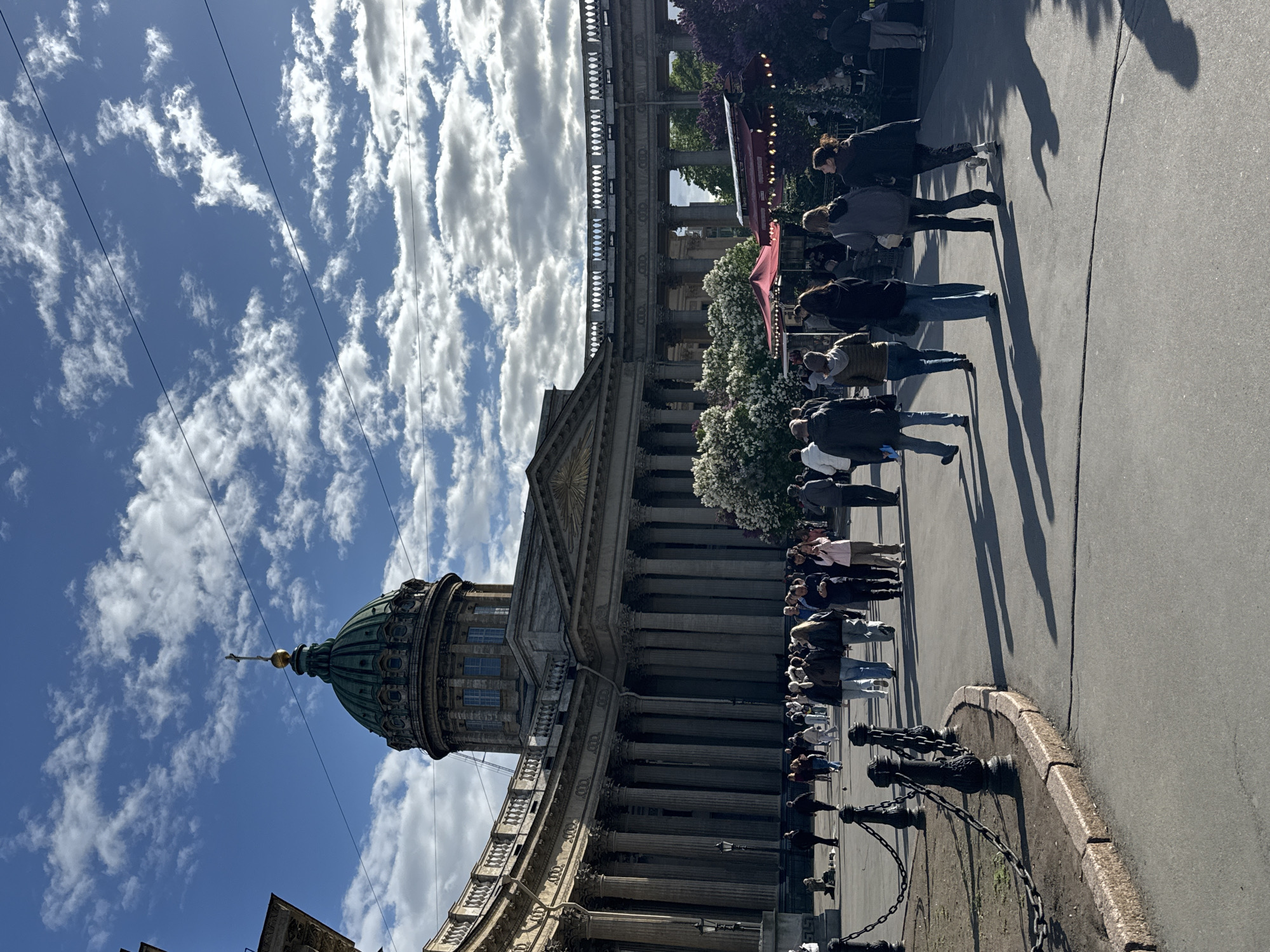 The image depicts a sunlit scene in Saint Petersburg, Russia, featuring the Kazan Cathedral. A crowd of people stand along the cathedral's side, likely waiting to enter. The cathedral's facade is visible, showcasing its neoclassical architectural style, including its dome, columns, and ornate detailing. An iron chain fence runs along the pavement. The sky above is bright blue with scattered white clouds. Utility wires cut across the upper part of the frame. The scene conveys a sense of a busy day in the city with tourists and locals visiting the landmark.