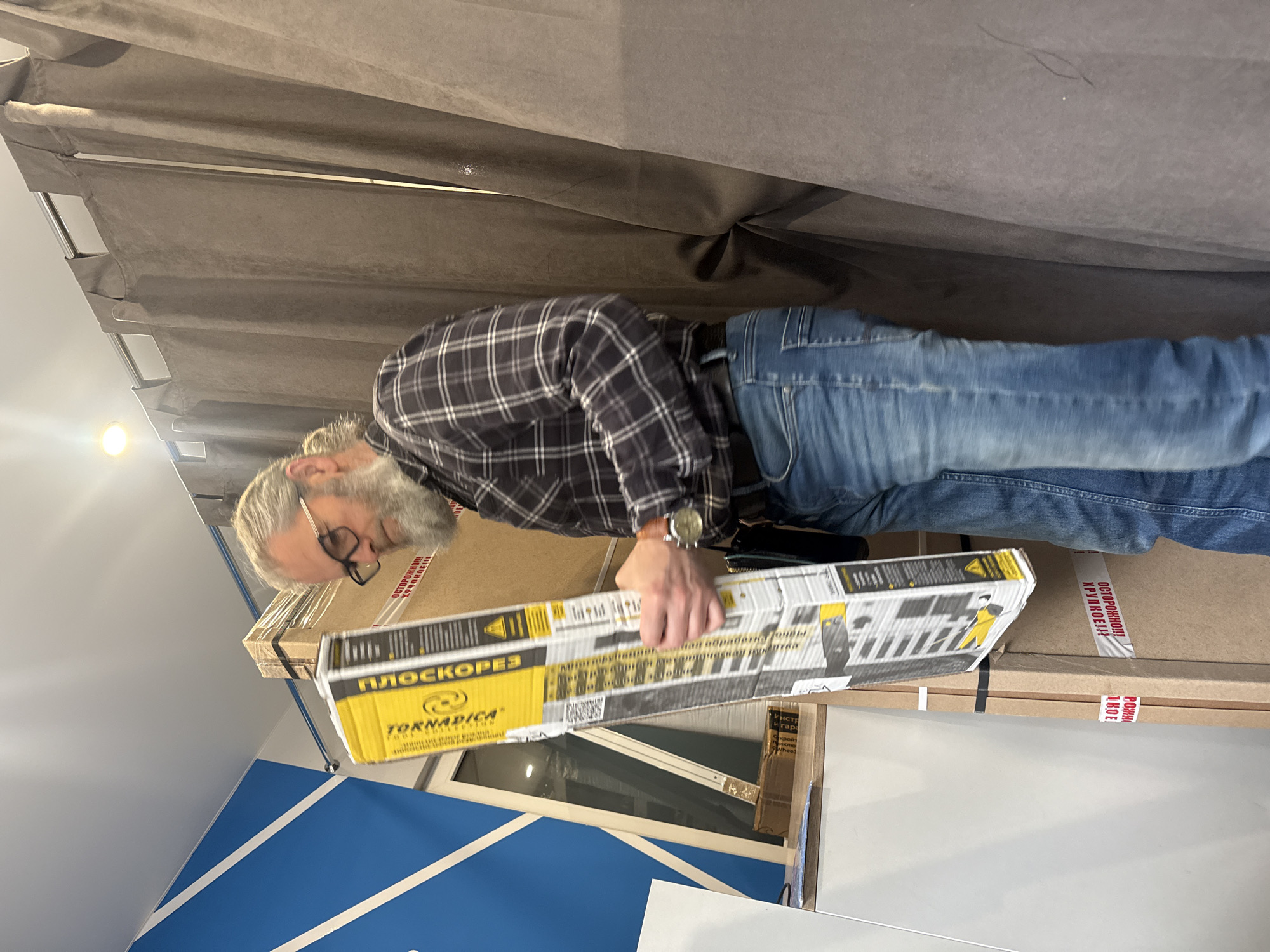 A man with grey hair and a beard is holding a yellow box with the word "ПЛОСКОРЕЗ" printed on it in what appears to be a room in Unknown, Unknown. He is wearing glasses, a checkered shirt, blue jeans, and a watch. The box is long and narrow with black and white graphics, and it sits atop other cardboard boxes. The environment features a large curtain in the background, a white wall with a blue geometric design, and a window.