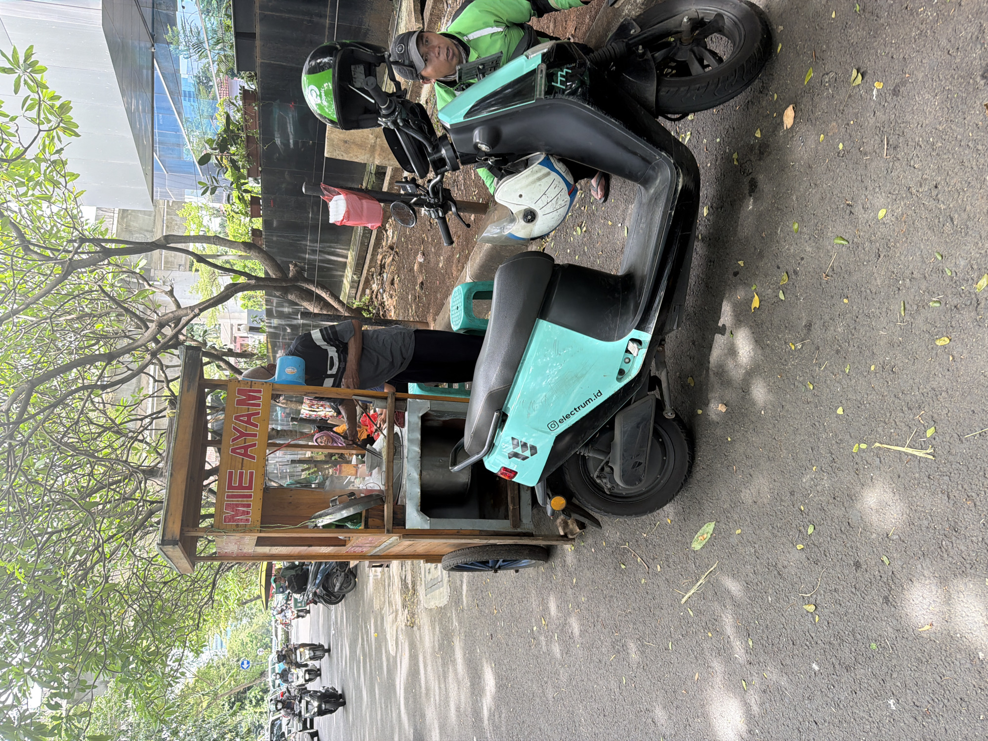 The image shows a light turquoise electric scooter with the writing "electrum.id" on its side, parked on a street in South Jakarta, Indonesia. A food cart with "MIE AYAM" written on its side is hitched to the scooter. A man in a green jacket and helmet is on a second scooter parked behind the electric scooter. More motorbikes can be seen parked on the street in the background. Buildings and trees are also visible. The street is paved and shows signs of wear and tear.