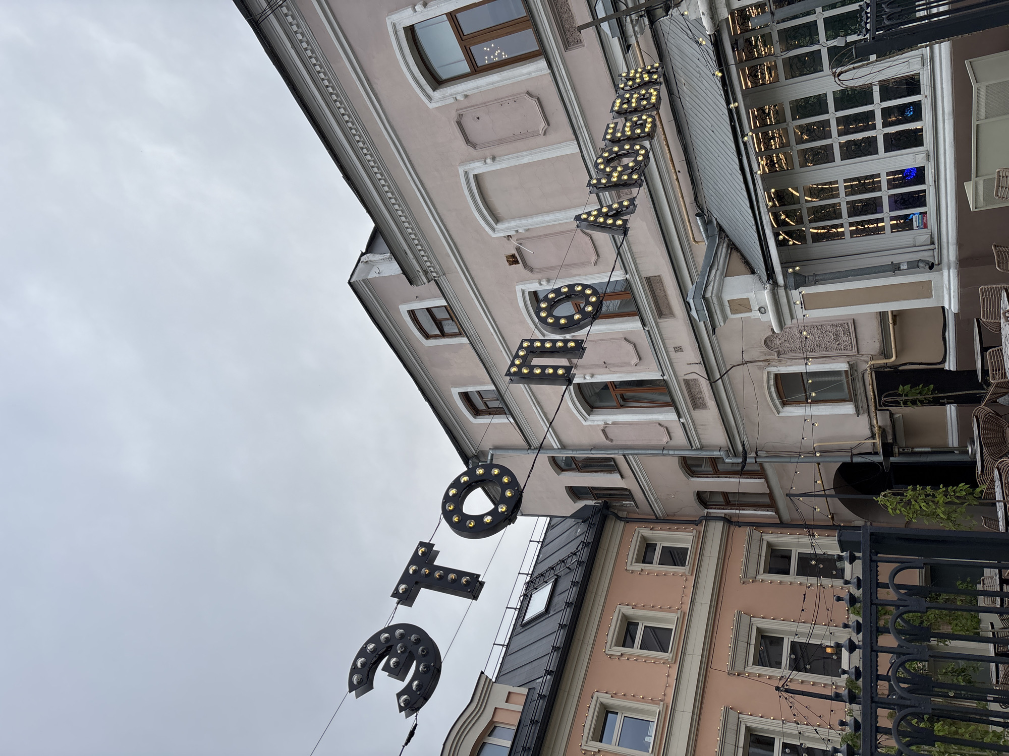 The image shows a street scene in Nizhny Novgorod, Russia, featuring two buildings with architectural details indicative of older European construction. Both buildings are painted in pastel colors, with one on the left being a lighter pink and the one on the right a slightly darker, muted pink. The buildings have multiple windows with dark frames, and the building on the right has decorative white trim around its windows and along the roofline.
Suspended across the street are large, illuminated letters forming a phrase in Russian. The letters are dark, three-dimensional, and outlined with numerous small, yellow lights. The phrase appears to read "ЭТО ЛЮБОВЬ" which translates to "This is love." The letters are strung on wires against a cloudy, overcast sky.
In the foreground, there is a black metal fence with decorative elements. Beyond the fence, outdoor seating with wicker chairs and tables is visible, suggesting a cafe or restaurant. String lights are also draped across the facade of the building on the left, adding to the festive or charming atmosphere. The overall impression is one of a quaint, possibly historic, urban environment, enhanced by the decorative lighting. There are no people visible in the image.