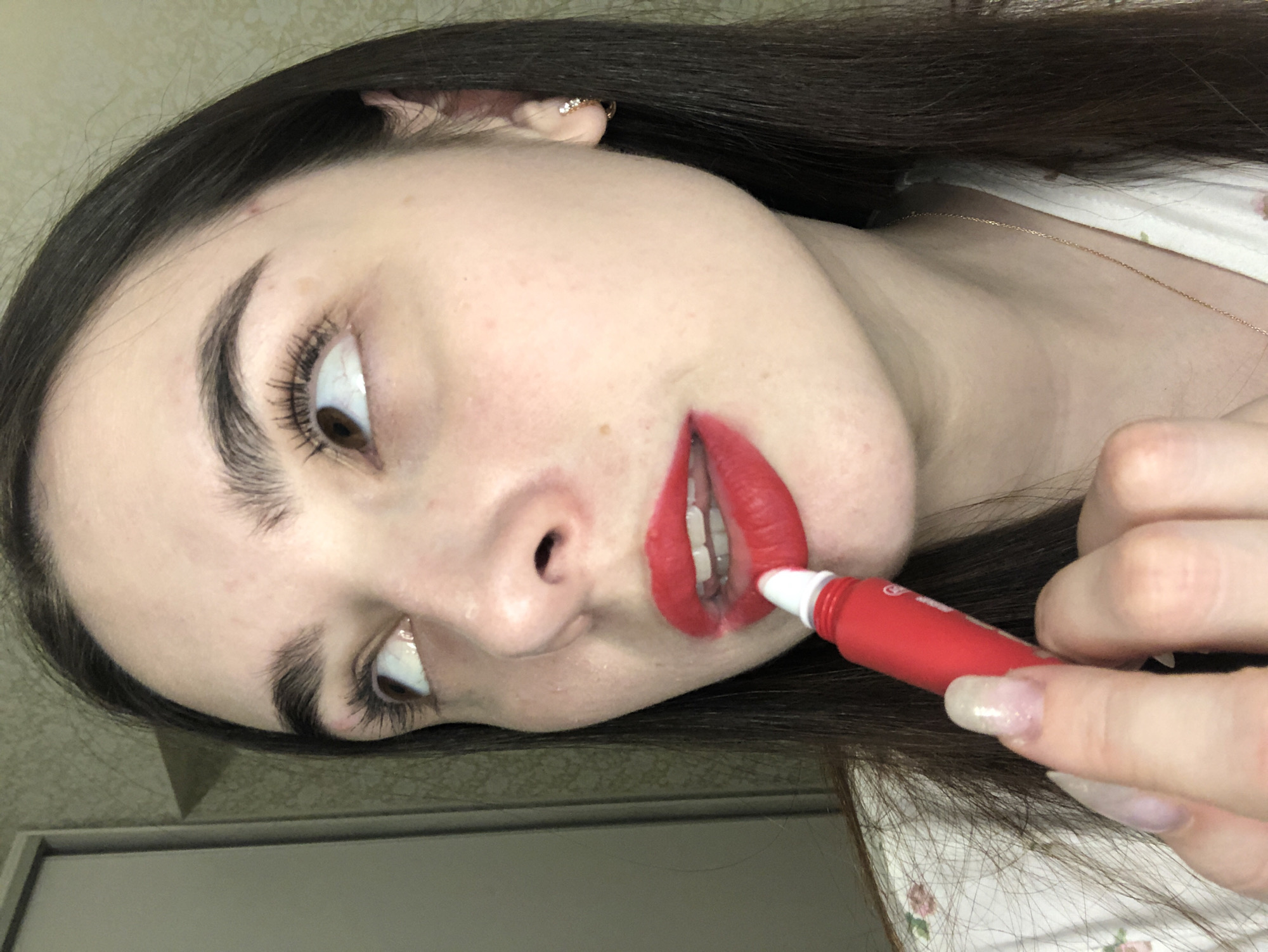 A woman with dark hair is applying red lipstick. She is wearing a light-colored floral top and a gold necklace. She has light skin, dark eyebrows, and brown eyes. She is holding a small red tube of lipstick in her right hand. The background appears to be a wall and part of a door frame. There is no indication of the city of Noviy, Russia, in the image.