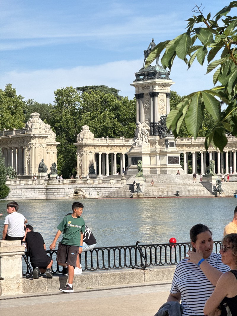 This outdoor image captures a vibrant scene at the Monument to Alfonso XII in El Retiro Park, Madrid, Spain, on a bright, sunny day. The dominant feature is the grand, classical-style monument with its elaborate columns, statues, and a prominent equestrian statue of King Alfonso XII atop a tall pedestal, situated across a large body of water.
In the foreground, several people are visible near a black metal railing that separates them from the water. On the far left, two young men are seated; one wears a white t-shirt, and the other a black t-shirt. Standing centrally, a young man in a green t-shirt and black shorts looks to his right. His t-shirt bears the text "All Time Legend." To the right, two women are engaged in conversation. One wears a striped t-shirt, and the other, with curly hair and glasses, touches the first woman's face, suggesting a close or affectionate interaction. A small red object, possibly a ball, is visible near the railing further back.
The water reflects the bright sky, creating a serene blue surface. In the background, beyond the monument, lush green trees fill the scene, indicating a large park setting. Numerous individuals can be seen walking on the steps and around the base of the monument, enjoying the sunny weather. The sky is clear blue with only a few faint wisps of clouds. The overall atmosphere appears relaxed and lively, typical of a popular public park on a pleasant day.
