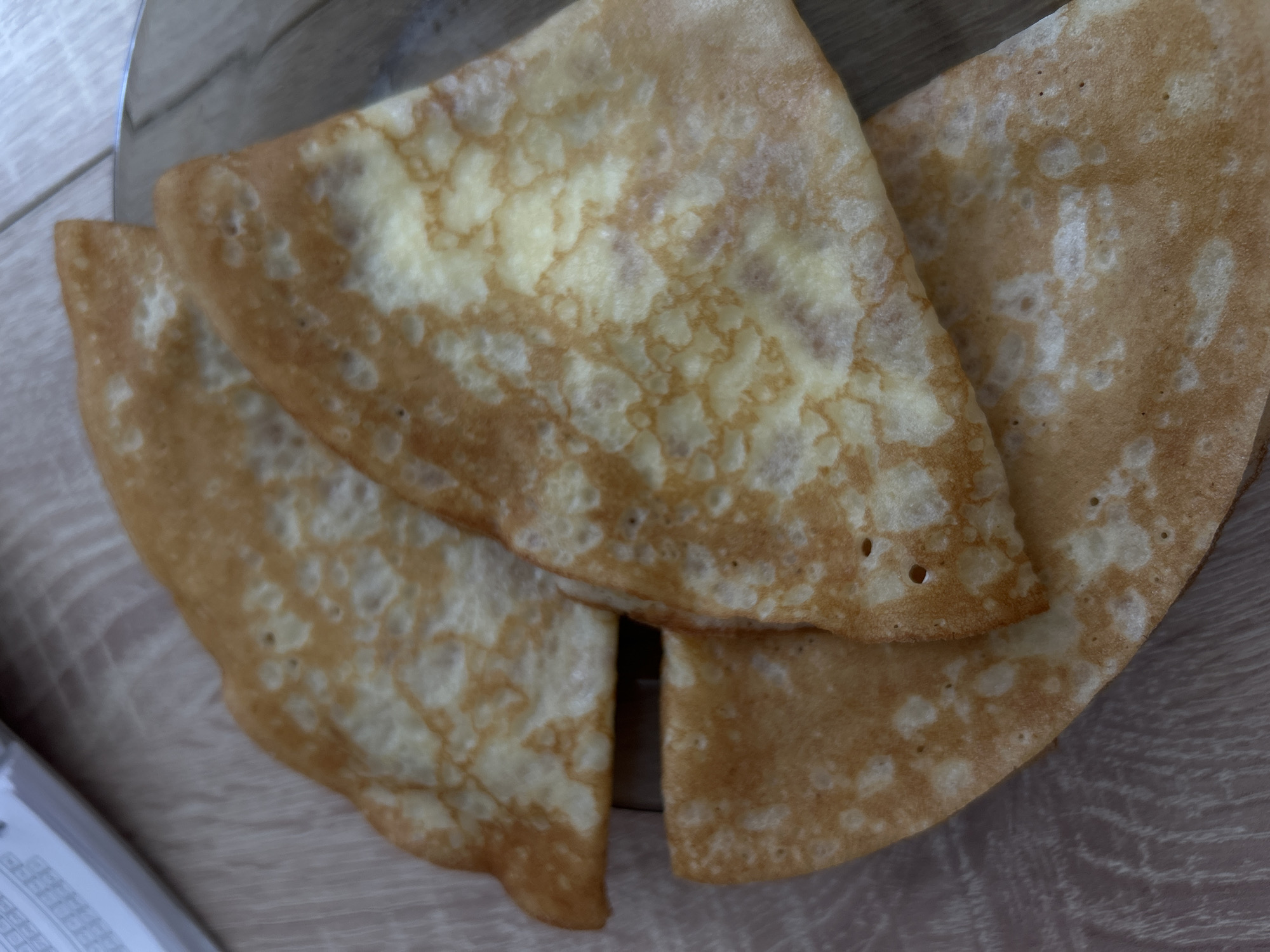 Three folded crepes, exhibiting uneven golden-brown and lighter yellow coloration consistent with pan-frying, are arranged on a light-colored wood-grain surface. One crepe occupies the bottom, with another partially overlapping it, and a third atop the stack, angled towards the upper right. The crepes show characteristic small bubbles and irregular browning patterns. In the upper-left quadrant, a portion of a white bound document or notebook with printed text is visible. A curved, reflective metallic edge, possibly from a plate or bowl, is present along the right side of the image. The scene is located in the city of Unknown, Unknown.