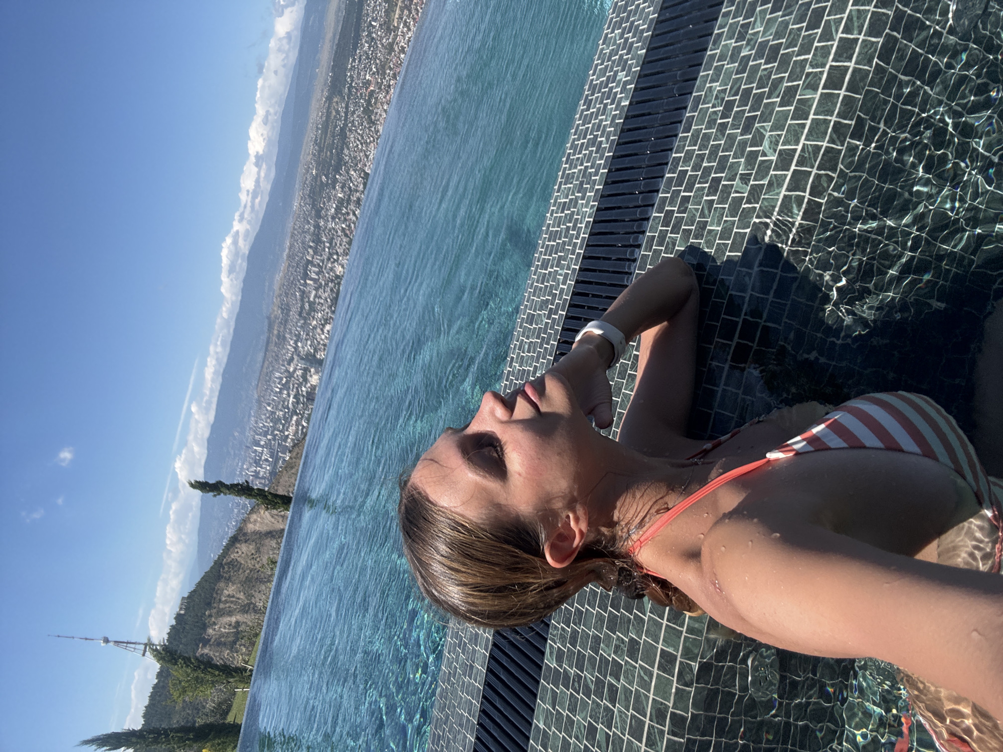 A young woman is captured in a selfie taken from a high vantage point overlooking a city. She is partially submerged in an infinity pool, her head resting on the edge. The pool's tiled edge, composed of dark green and white mosaic tiles, contrasts with the bright blue water. Beyond the pool, a sprawling cityscape stretches out, nestled within a valley and bordered by distant mountains. The sky is a clear, vibrant blue with a few wispy clouds. The woman is wearing a striped bikini. Her skin is wet, and water droplets are visible. She appears relaxed and content, her eyes closed as she basks in the sunlight. The scene suggests a warm, sunny day, possibly in the late morning or early afternoon, given the bright light and clear sky. The presence of a prominent television tower on a hill in the background, a known landmark in Tbilisi, Georgia, helps to identify the location.