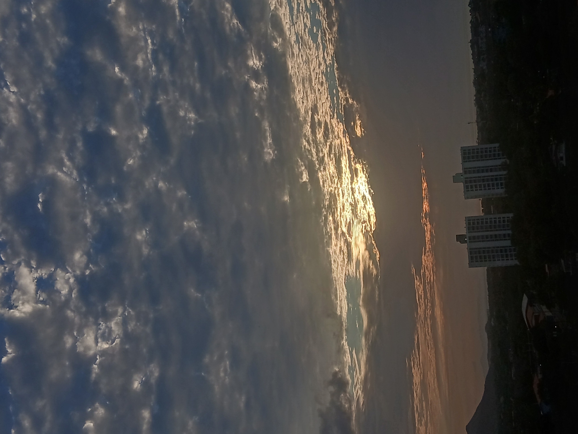 The image shows a sunset over the city of Juan Griego, Venezuela. The sky is filled with clouds, with the sun shining through a gap in the clouds, creating a bright, golden effect. Buildings are visible on the right side of the image, and a mountain or hill is in the background. The image appears to be taken from an elevated position.