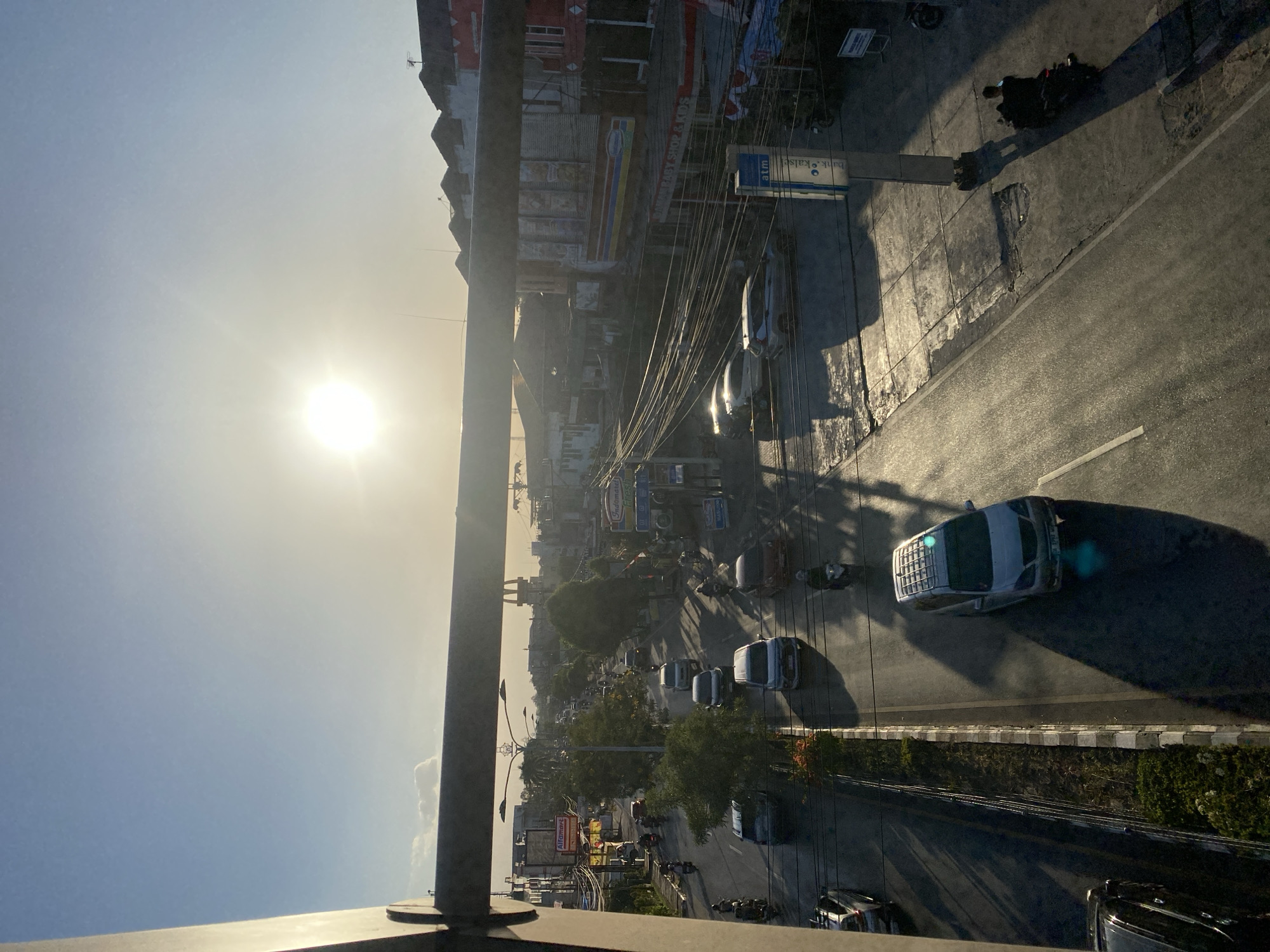 The image captures a street scene in Banjarbaru, Indonesia, during what appears to be sunset, given the low sun angle and warm illumination. A prominent horizontal pipe or beam crosses the upper portion of the frame, casting a shadow. Below it, a two-lane road is visible, with traffic consisting of cars and motorcycles. Some vehicles are moving, while others are parked. On the right side of the road, there are buildings, some with awnings and signs, including what looks like an ATM and a store. A lamppost with two lights is present, and numerous electrical wires crisscross overhead, typical of urban infrastructure. The road surface shows reflections from the sun, indicating it might be wet or have a glossy finish. The general environment suggests a commercial or mixed-use urban area.