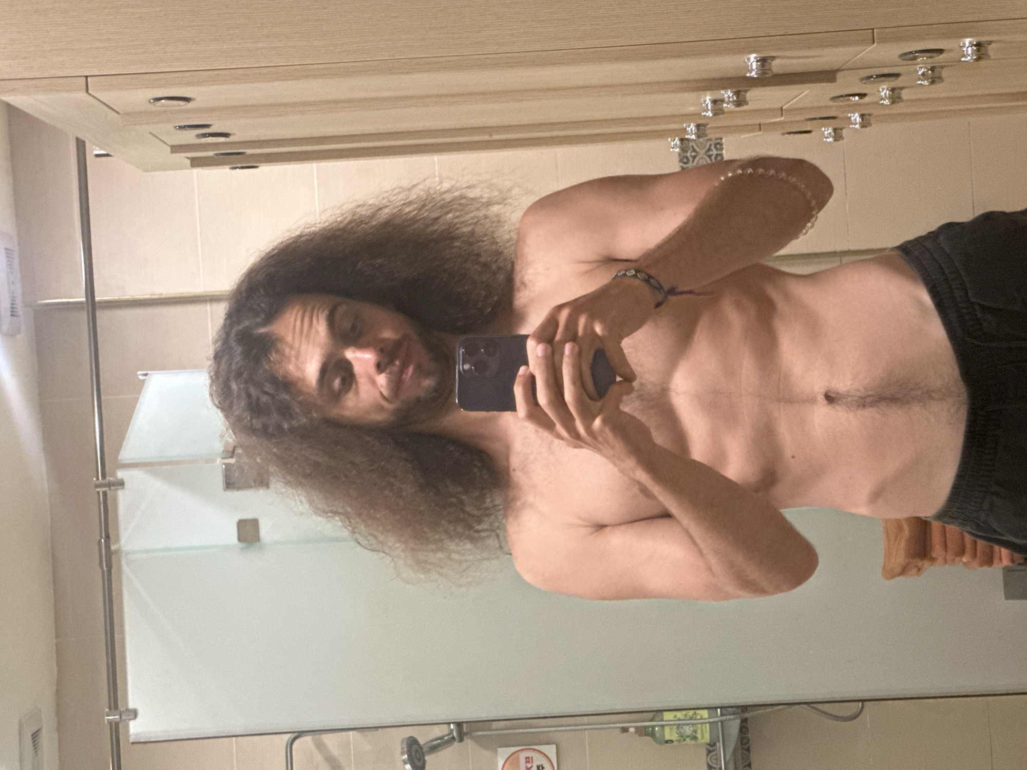 The media file is a mirror selfie of a shirtless man with long, curly brown hair. He is wearing dark shorts and has a beaded bracelet on his left wrist. The man is holding a smartphone in his right hand, angled to capture the reflection. His expression is relaxed and he appears to be in a bathroom.
The setting suggests a private space, likely a bathroom, with tiled walls and a wooden vanity with several cabinet knobs visible. A frosted glass partition is present in the background, hinting at a shower or bathing area. The lighting is artificial and appears to be from overhead, suggesting it is either daytime or nighttime indoors. There are no clear indications of the weather. The reflection in the mirror shows the man from the waist up.
The activity is a selfie being taken by the man. There are no explicit emotional cues beyond a relaxed demeanor. No text is clearly visible or decipherable in the image. The location context provided is Đà Nẵng, Vietnam, but there are no specific visual elements within the image that confirm this location.