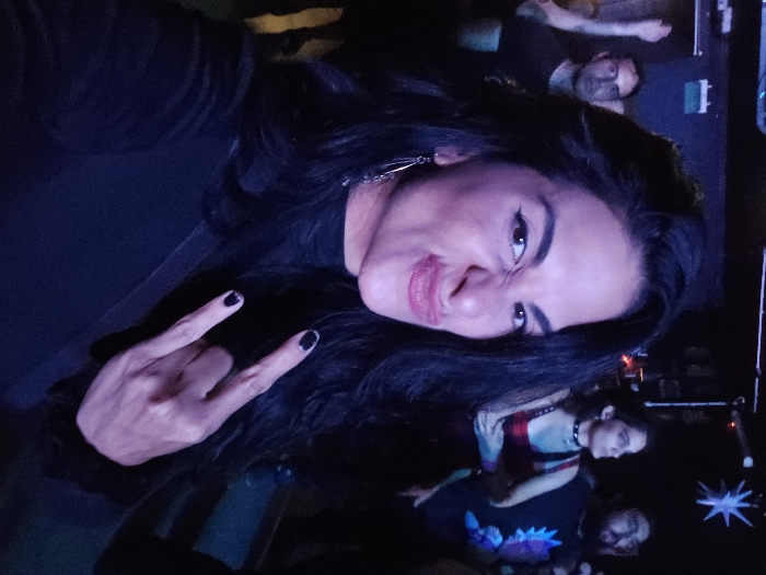 This image depicts a woman captured in a selfie within a dimly lit indoor live music venue in Buenos Aires, Argentina. The central subject, a woman with long dark hair, presents a facial expression characterized by pursed lips and a wrinkled nose. She wears a black long-sleeved top and silver-colored dangling earrings. Her right hand, adorned with dark nail polish, performs the "rock-on" gesture. The background, illuminated primarily by blue and purple artificial lighting, shows a crowd of attendees. To the left, a man in a dark shirt is visible with his left arm raised. To the right, a woman wearing a choker necklace and a man with a beard are present, both looking towards the left side of the frame. Overhead rigging, a white star-shaped decorative light, and a green sign featuring a white numeral "1" are discernible in the environment.