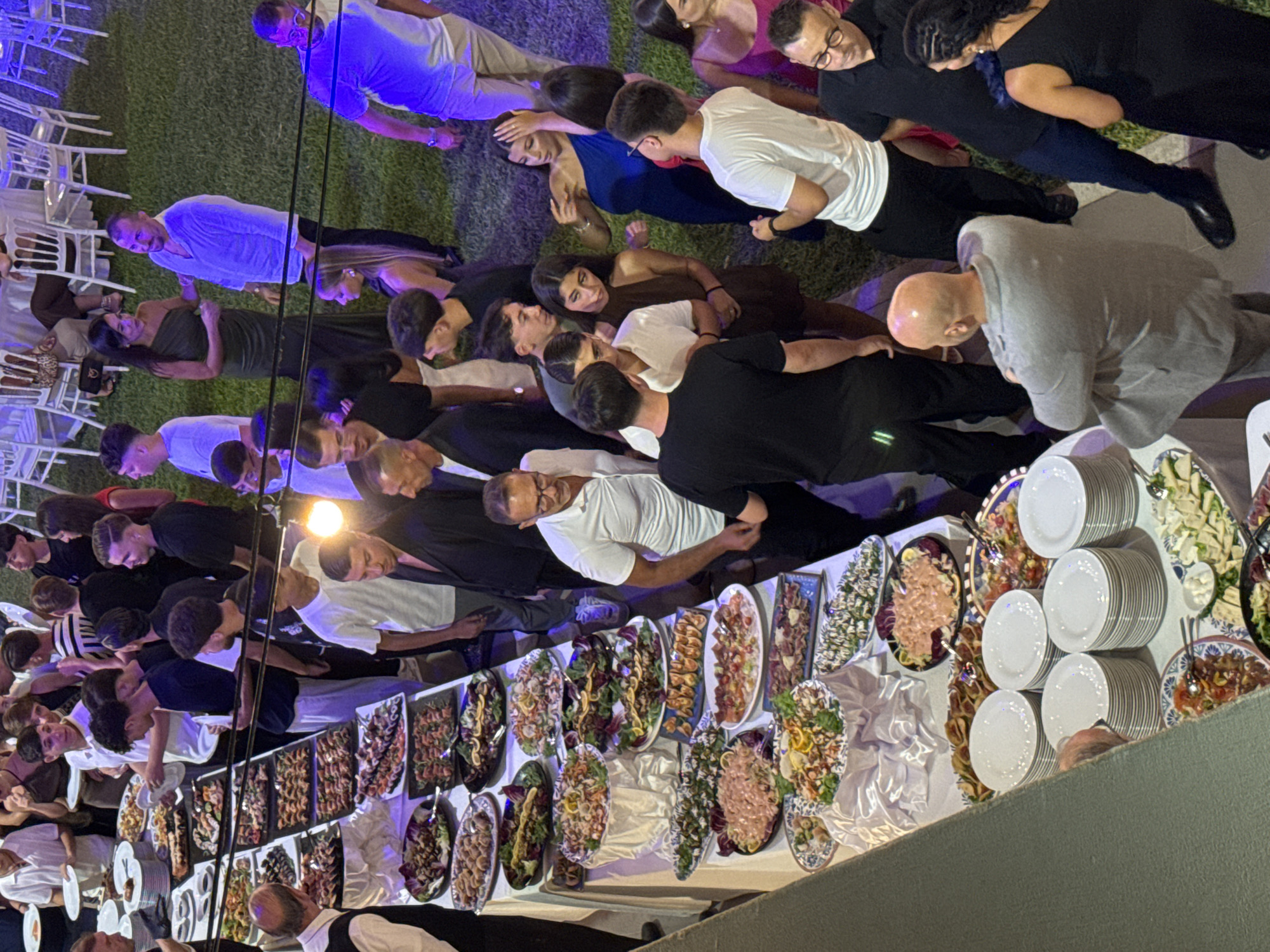 This high-angle outdoor photograph captures a lively scene at a catered event, likely a wedding reception, set against a grassy backdrop. The event appears to be taking place in Isola delle Femmine, Italy, as suggested by the presence of a buffet setup with various seafood and meat dishes, a common feature at Italian celebrations.
The foreground is dominated by a long buffet table laden with an abundance of food. Platters of seafood salads, cured meats, antipasti, and other prepared dishes are artfully arranged, attracting the attention of the guests. Several stacks of white plates are neatly placed at the front of the buffet, ready for guests to serve themselves.
In the midground and background, numerous people are milling around, engaged in conversation and enjoying the festivities. They are dressed in a mix of casual and formal attire, with men in shirts and trousers and women in dresses. The lighting suggests it is evening, with some artificial lights illuminating the scene. Several tables with white tablecloths and chairs are visible in the background, hinting at dining areas.
The overall atmosphere is one of a festive gathering, with people interacting and enjoying the food and company. The lush green grass surrounding the event space adds to the idyllic setting.