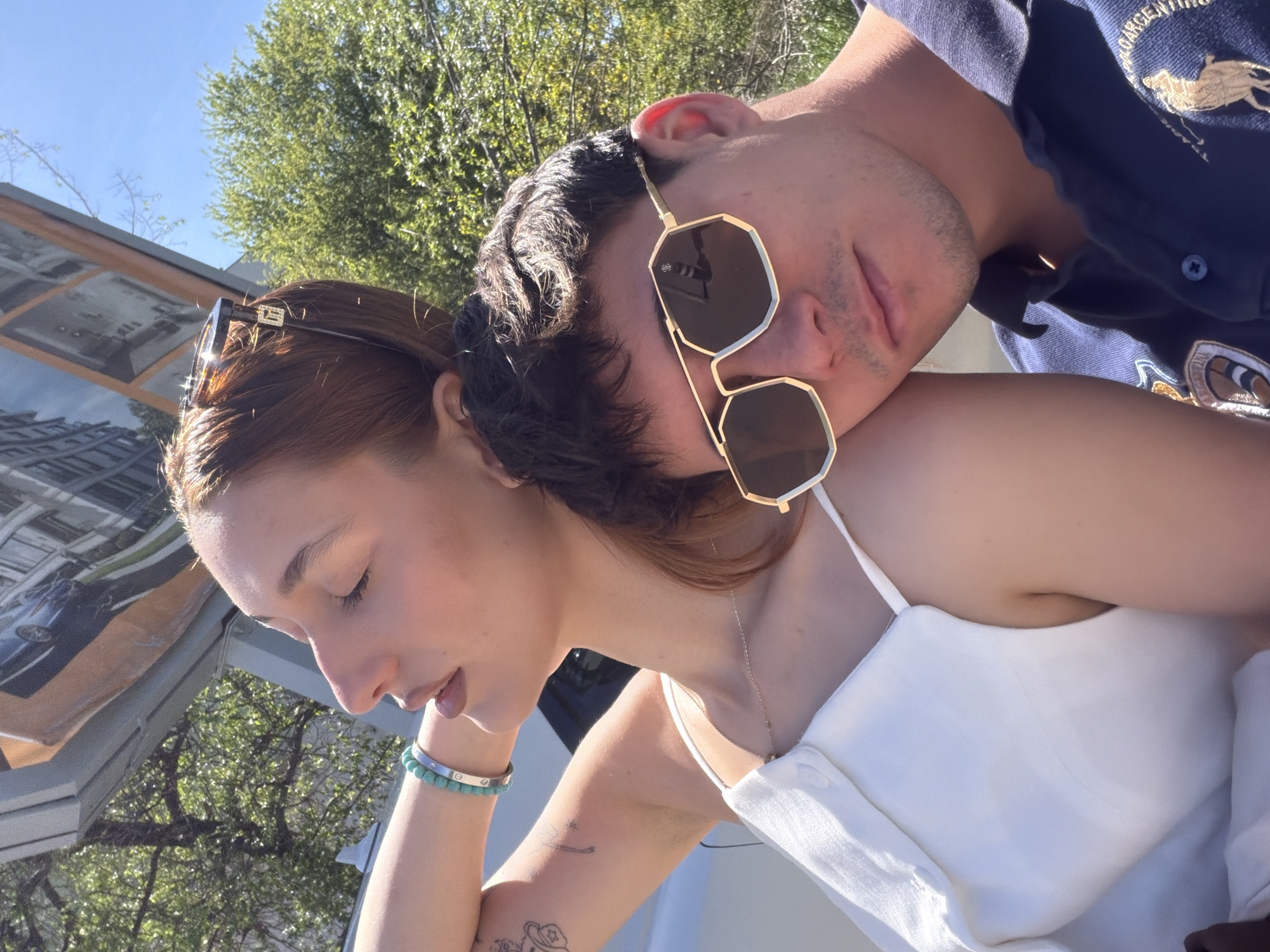 A young couple is pictured in an outdoor setting, possibly in Sofia, Bulgaria, although specific landmarks are not visible. The woman has auburn hair and is wearing a white tank top and a bracelet. A man is positioned with his head resting on her shoulder, and he is wearing sunglasses and a navy blue polo shirt. The background includes trees and a structure with reflective glass. The photo is taken in daylight.