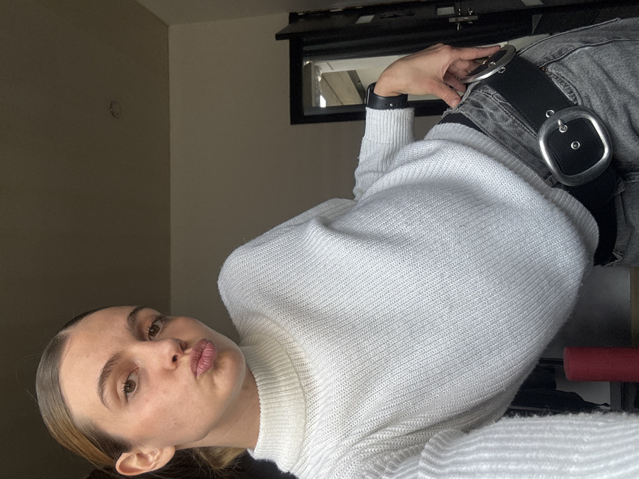 The scene is an indoor setting in Tbilisi, Georgia, illuminated by natural light from an unseen source to the right, creating a warm ambiance.
A young adult female is centered in the frame, angled towards the left, taking a selfie. She has light-toned skin with visible minor imperfections and light brown hair pulled back. Her lips are pursed in a "kiss" pose, and her brown eyes are directed at the camera.
She is dressed in a white or off-white knitted turtleneck sweater with horizontal ribbing, and grey, possibly distressed, denim jeans. A black belt with two prominent silver-toned, oval-shaped buckles is worn over her jeans. On her left wrist, she wears a black cuff, possibly a smart device or watch, with two small studs visible. Her left hand is positioned near her hip, touching the lower buckle of her belt.
The background consists of a plain, light-colored wall, likely beige or off-white, with a ceiling line visible. In the upper right, a small, circular recessed light fixture is visible on the ceiling. To the right of the subject, a dark-colored door frame and a partially open, dark-colored door are visible. Beyond the door, a glimpse of another interior space, possibly a hallway or stairs, is present. In the lower-left corner, a dark red cylindrical object, possibly a yoga roller, is partially visible against the wall.