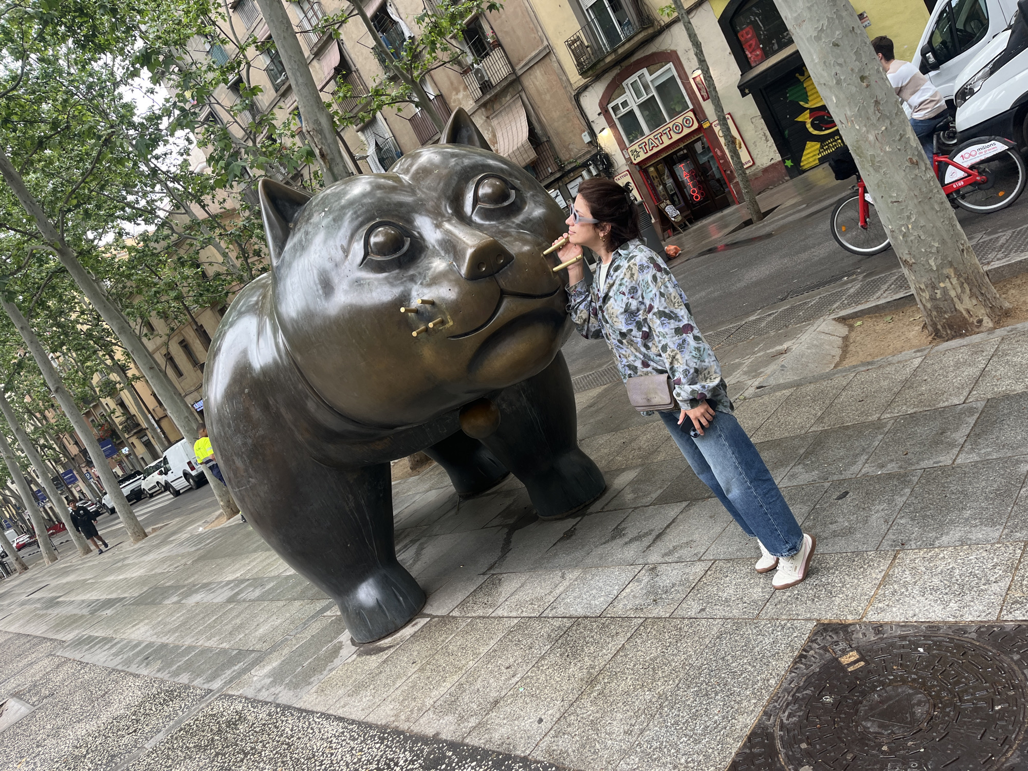 Here's a detailed description of the image:
**General Overview:**
The image captures a street scene, likely in Barcelona, Spain, featuring a large bronze cat sculpture. A woman is interacting with the sculpture.
**People:**
* A woman is standing next to the cat sculpture. She has dark hair pulled back, sunglasses, a floral-patterned long-sleeved shirt, blue jeans, and white sneakers. She is holding two wooden sticks to the cat's face, seemingly 'giving it a mustache'.
* In the background, a person is riding a red and white Bicing bike.
* Other people are visible walking along the street.
**Objects:**
* **Cat Sculpture:** A large, bronze sculpture of a cat is the central focus. It is depicted in a squatting position.
* **Wooden Sticks:** Two wooden sticks are visible being held up to the cat's face.
* **Bike:** A red and white Bicing bike is visible in the background.
* **Buildings:** The background features buildings, including one with "TATTOO" written on the storefront.
* **Trees:** The street is lined with tall trees.
* **Street Furniture:** A manhole cover is visible in the foreground.
* **Vehicles:** There are cars and a white van in the background.
**Actions and Interactions:**
* The woman is posing with the cat statue, holding the wooden sticks up to the cat's face.
* People are walking and biking in the background.
**Environment:**
* **Location:** The scene appears to be a street in a city, likely Barcelona, Spain. This is indicated by the architectural style, the "TATTOO" signage (common in tourist areas), and the Bicing bike.
* **Streetscape:** The street has a tiled sidewalk.
* **Weather:** The weather appears to be overcast or partly cloudy.
* **Time of Day:** It appears to be daytime.
* **Landscaping:** Trees are planted along the street.