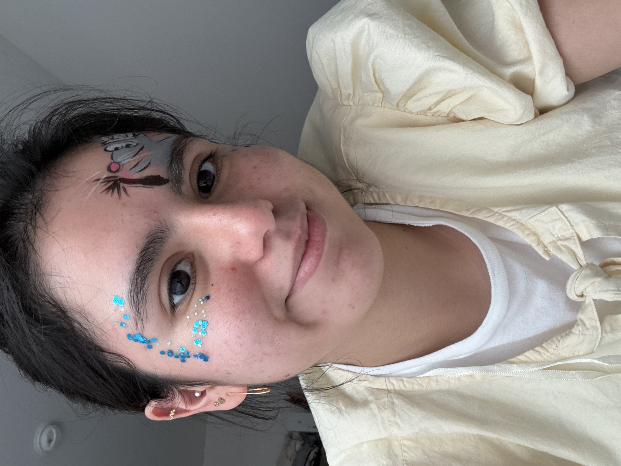 In this close-up, eye-level selfie, a young woman with dark hair and fair skin is featured. She has blue glitter applied to her forehead and under her right eye, and a cartoonish drawing of a cat-like creature with pink cheeks and wide eyes is painted on her forehead. She is wearing a light yellow, puff-sleeved top over a white t-shirt, and gold earrings. The background is a plain, light grey wall. The image was captured indoors, with soft lighting. The location is not specified in the image, and therefore cannot be determined to be Austin, United States of America.