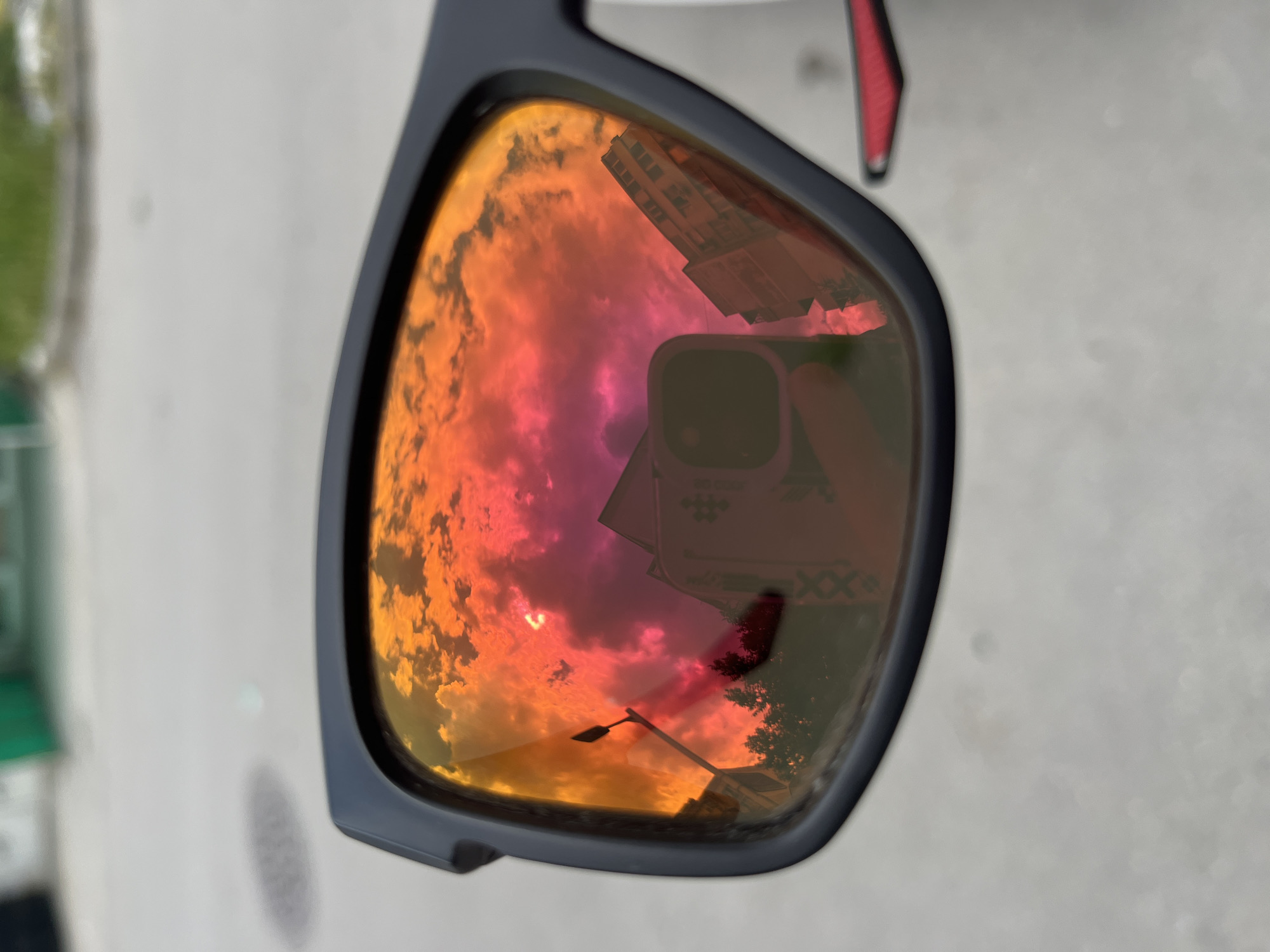 A pair of sunglasses reflects a vibrant sunset sky, a tall building, and a cell phone held by the photographer. The scene captured in the reflection suggests an urban setting. The presence of the building and general urban atmosphere hints at a location possibly within a city such as Varna, Bulgaria, known for its coastal cityscape. The cell phone is held up to capture the reflection in the sunglasses, making the photographer a subject of the image. Other details are the lamp post, and the building with roman numerals.