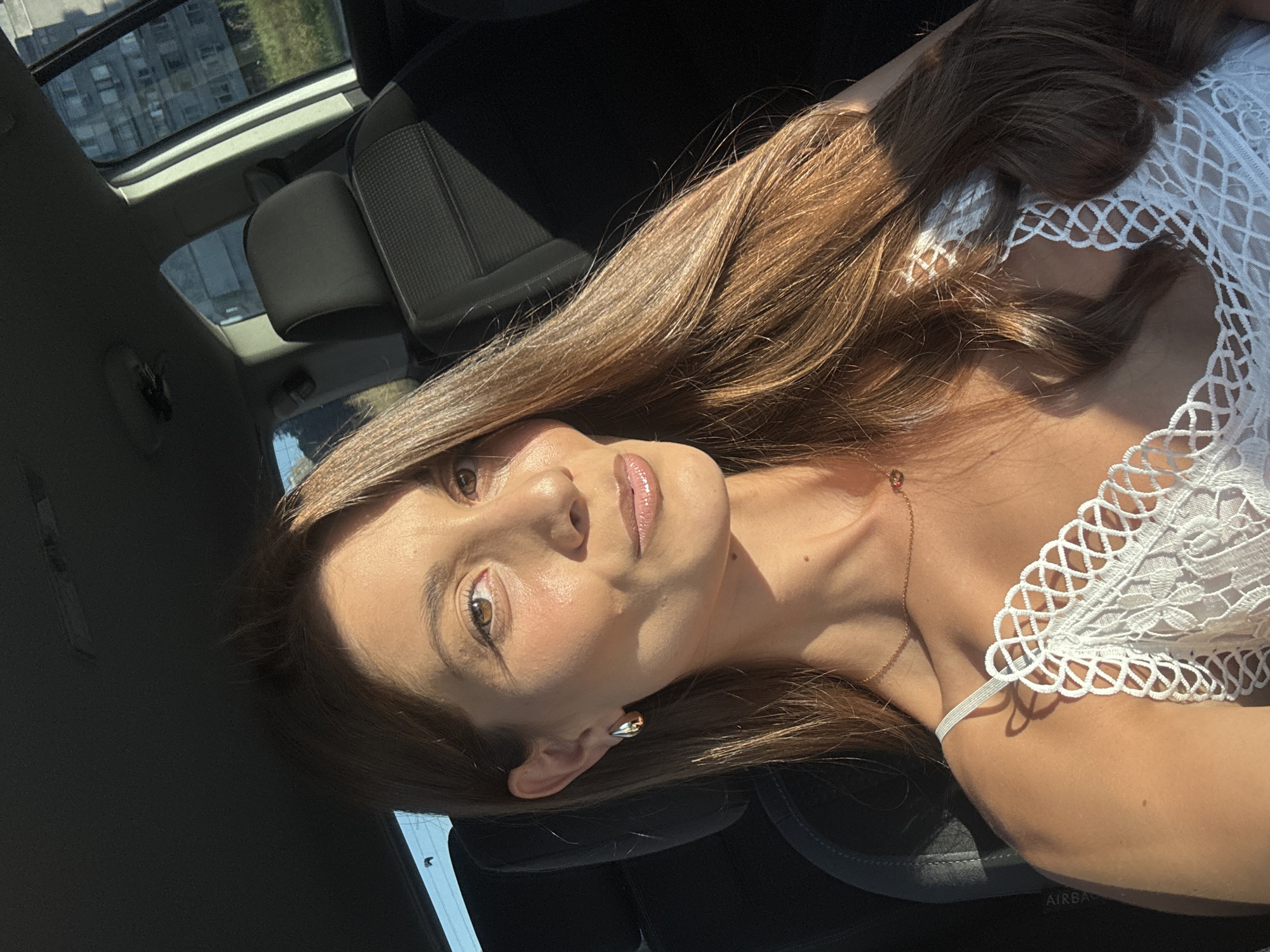 A woman with long brown hair is pictured in the driver's seat of a car. She is wearing a white, lacy top and gold earrings. The sun is shining through the car windows, casting shadows. Outside the car, a block of apartment buildings and some trees are visible, consistent with an urban setting in Burgas, Bulgaria. The image is a selfie taken from inside the vehicle.