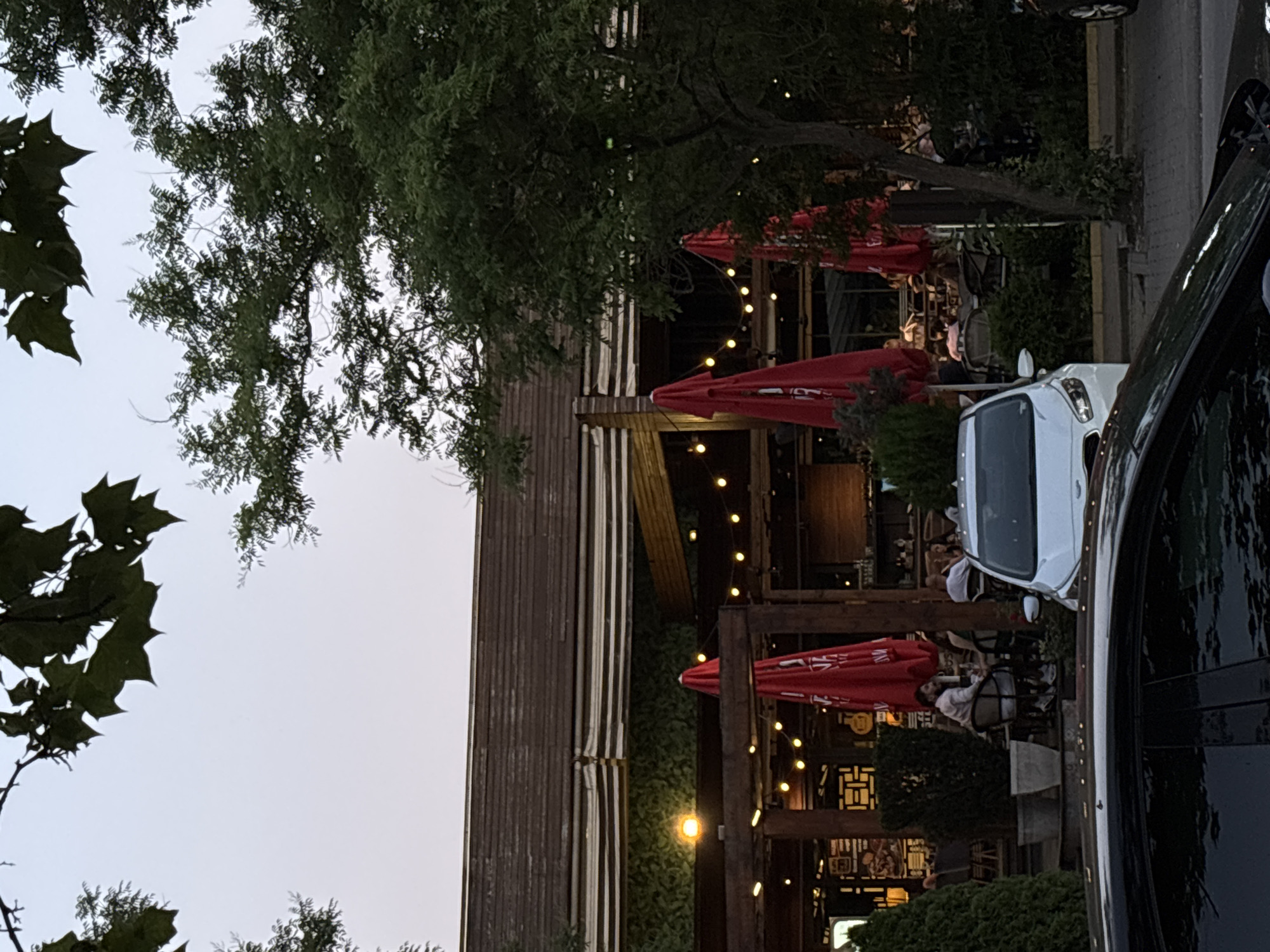 This is a photograph of an outdoor dining area at a restaurant, possibly in the evening as evidenced by the string lights. The scene is busy with people seated at tables, enjoying meals and conversations. Red umbrellas provide shade over some of the tables. A white van is parked near the dining area. The restaurant appears to have a rustic, wooden design, with vertical wooden planks on one of the structures. Trees with green foliage are visible, suggesting a natural or park-like setting. The sky is overcast and light. There is no visible text in the image.