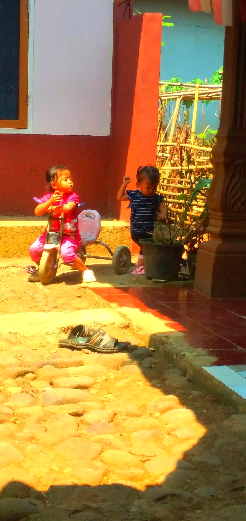 Two young children are in an outdoor area. The child on the left, wearing a pink outfit, sits on a pink and blue child's tricycle. The child on the right, wearing a striped shirt, stands and appears to be interacting with a potted plant. The ground is covered in sand and rocks, with some red tiles visible in the lower right corner. In the background, there is a building with red and white sections, and a bamboo structure. This scene is located in Rahong, Indonesia, in what appears to be a residential or yard area.
