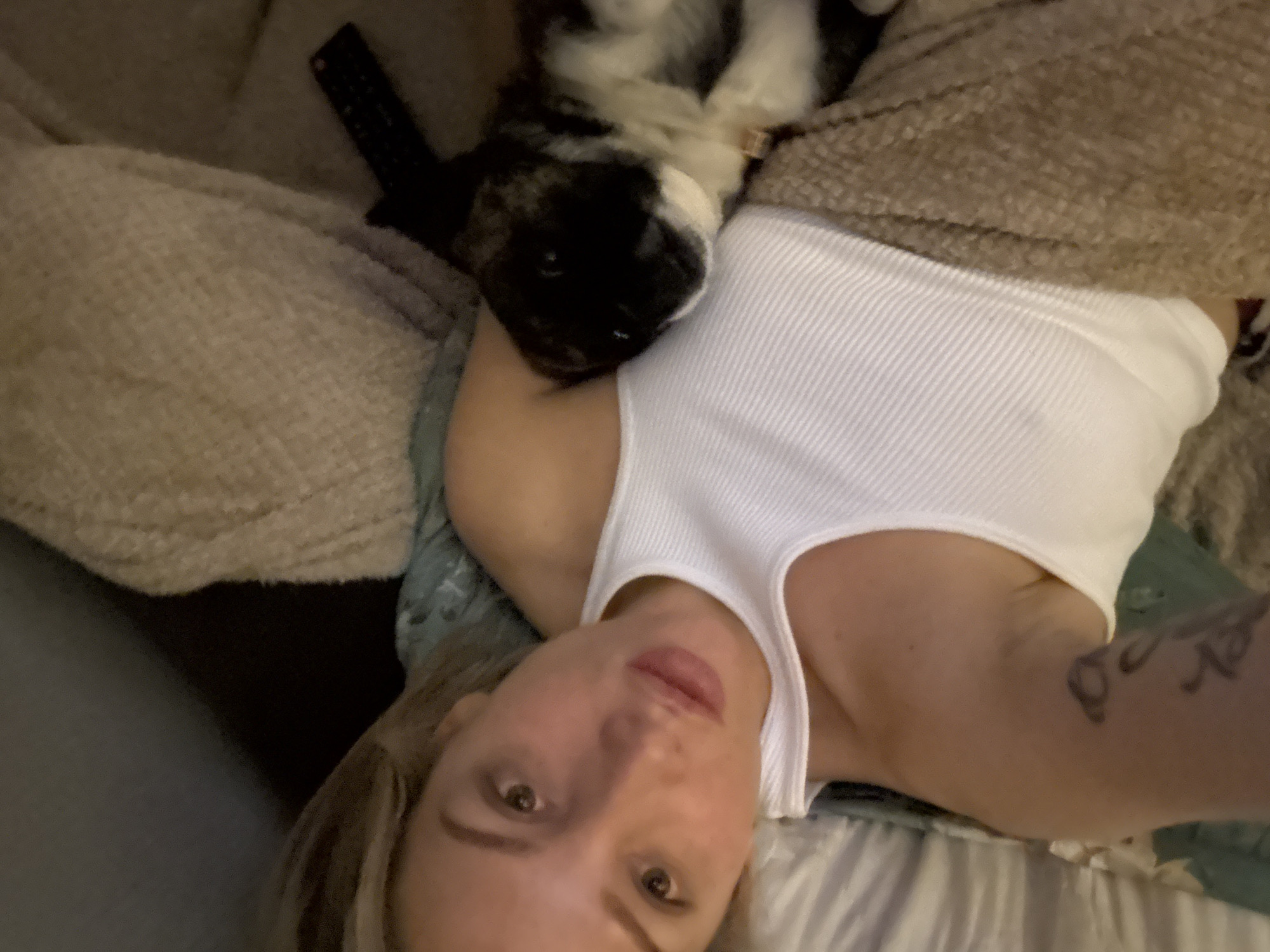 A selfie-style shot depicts a young woman and a small, fluffy dog resting together indoors in dim lighting, suggesting evening or night. The woman, with light brown hair, is lying down and looking towards the camera with a neutral expression. She wears a white ribbed tank top. A black and white puppy or small dog is nestled on her left shoulder and chest, appearing calm.
The setting is an interior space, likely a bed or couch, with soft textures from blankets and pillows. A light brown, textured blanket covers parts of the scene, including the woman's right side. To the upper left, a dark remote control is visible on a light-colored, textured surface. A tattoo with dark, stylized, illegible lettering is partially visible on the woman's left arm. The overall atmosphere conveys relaxation and companionship. The location context for the image is Moscow, Russia.