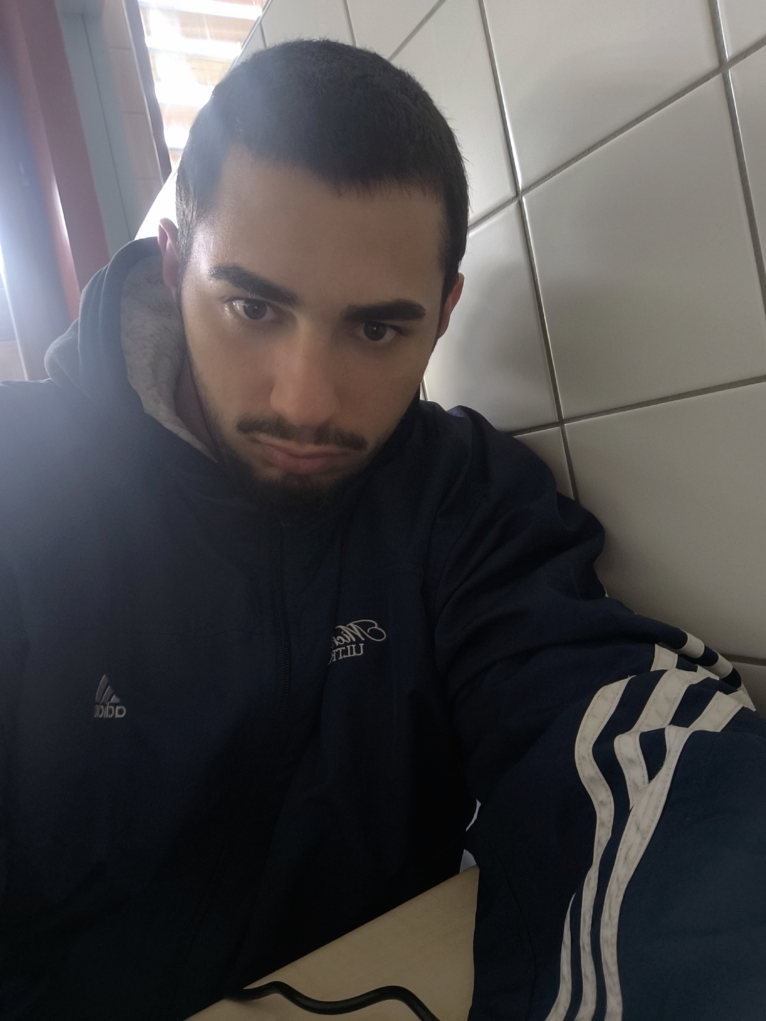 A man with short dark hair, dark eyebrows, and a beard is wearing a dark blue jacket with white stripes on the sleeve and a gray hoodie underneath. He appears to be indoors, sitting next to a wall of white tiles and a window covered with a red and white patterned curtain. The location is potentially in Schiltigheim, France.