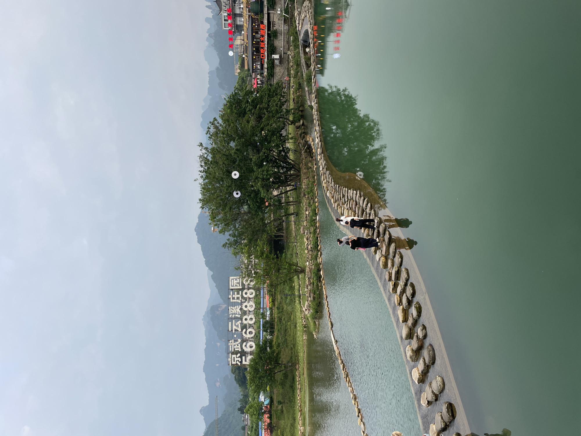The image shows a landscape in Wulingyuan, China. Two people are walking on a path made of stone that crosses a body of water. A tree and a grassy area are adjacent to the path. A sign with Chinese characters and the number "666888" is visible on the other side of the water. Mountains are in the background. The sky is overcast.
