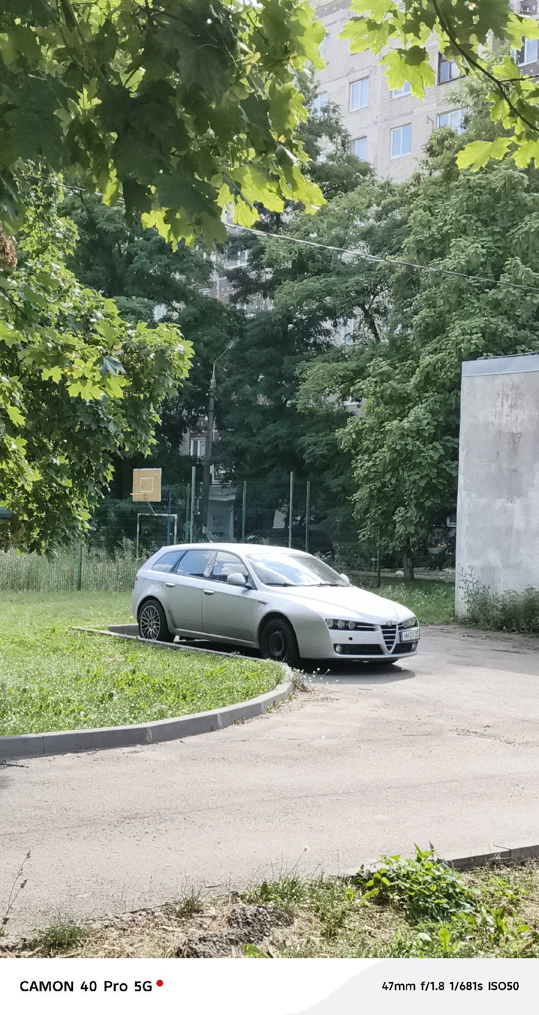 In Kramatorsk, Ukraine, a silver Alfa Romeo station wagon is parked on a paved area next to a grassy lawn. The car, facing left, has its front wheels turned slightly. Behind the car, a basketball hoop with a yellow backboard is visible, along with a chain-link fence. Lush green trees surround the scene, and a multi-story building with numerous windows is in the background. Sunlight filters through the leaves of a large tree in the foreground, casting shadows. The image captures a daytime scene in an outdoor urban environment.