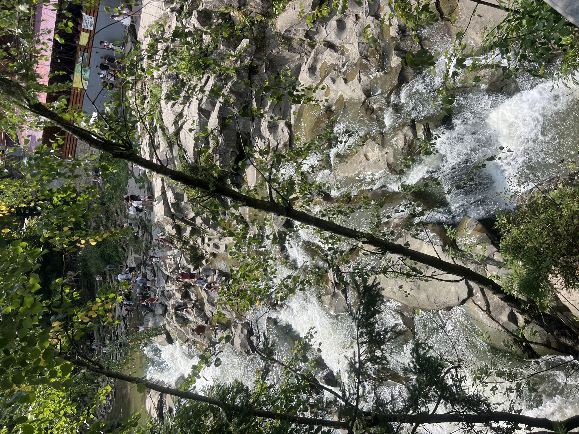 This image depicts the Prut River flowing through a rocky gorge in Yaremche, Ukraine. Several people are visible on the rocks near the river, some appearing to be bathing or wading in the water. The river cascades over numerous rocks, forming small waterfalls and rapids. Lush green foliage, including trees and bushes, frames the scene from the foreground and surrounding banks. In the background, a building with a red roof and signs is visible, suggesting a commercial area or restaurant. The environment appears to be a natural attraction with a developed tourist infrastructure.