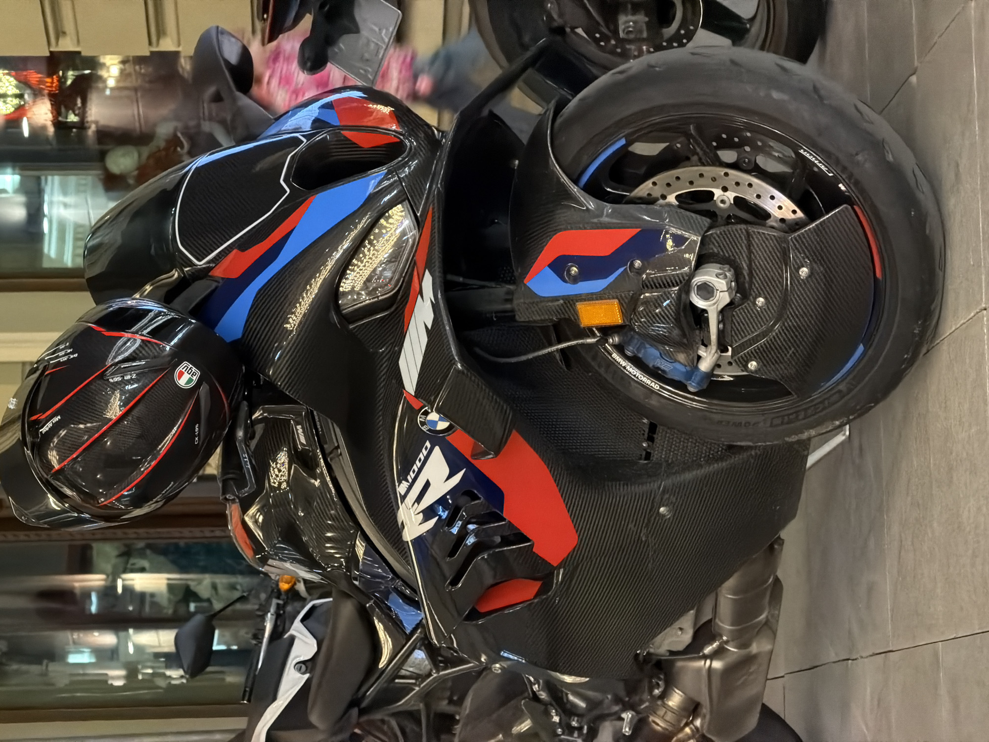 The image shows a BMW M 1000 RR motorcycle, characterized by its carbon fiber bodywork and the distinctive blue, red, and white BMW Motorsport color scheme. A black AGV helmet with red accents hangs on the left handlebar. The motorcycle is indoors, likely in a showroom or dealership. The background is blurry, but it appears to be a retail environment with people and displays visible through a glass partition. The precise location in Moscow, Russia, is not discernible from the image.